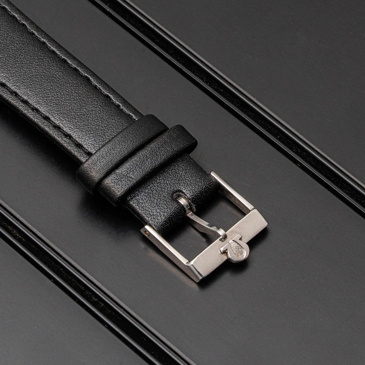 A close-up of a black leather watch strap with a silver-toned metal buckle, laid diagonally on a dark textured background.