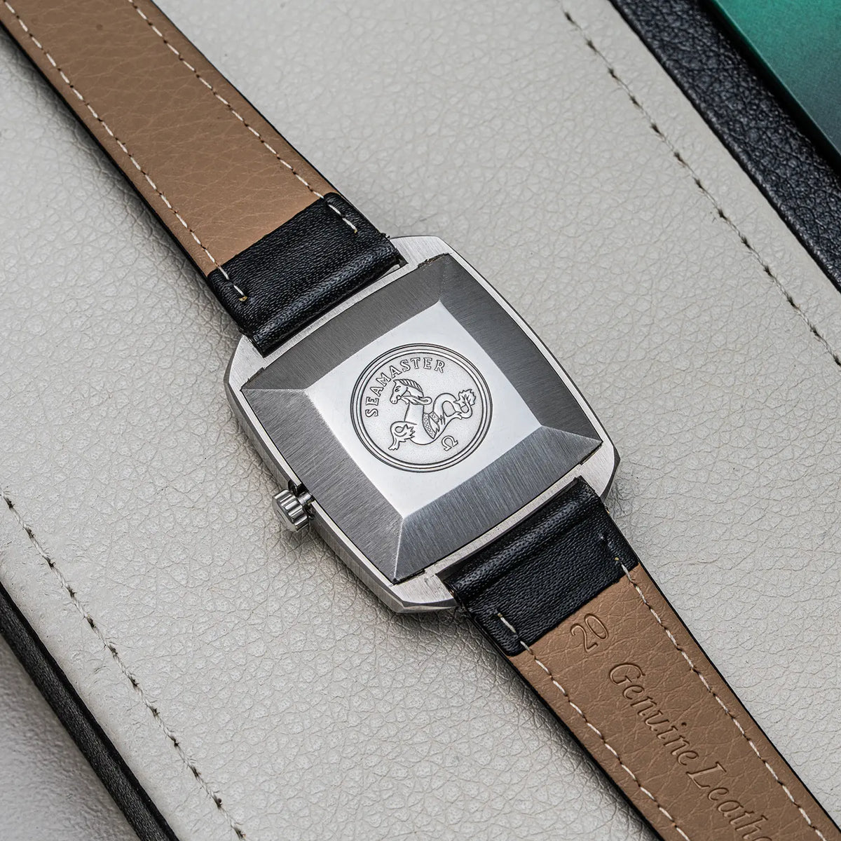 Close-up of a wristwatch Omega Seamaster, Ref. 166.042, Cal. 565, Men’s Automatic back showing a brushed silver-tone case and a tan leather strap with black edges, resting on a light textured surface.