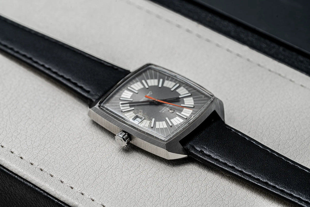 A close-up of a rectangular analog watch with a silver-toned metal case, a dark gray dial, white hour markers, and an orange second hand, resting on a light gray textured leather surface.