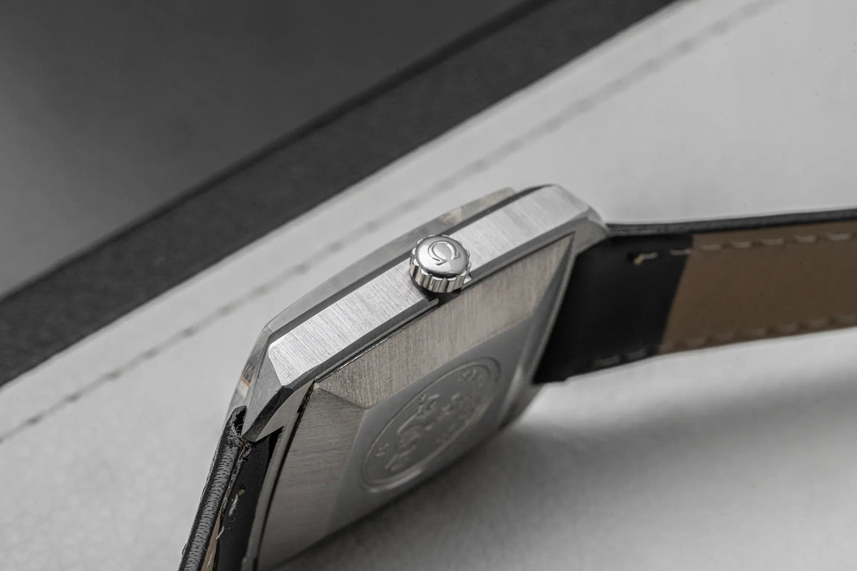 Close-up of a stainless steel watch case showing the brushed metal finish, crown, and part of a dark leather strap on a light gray surface.