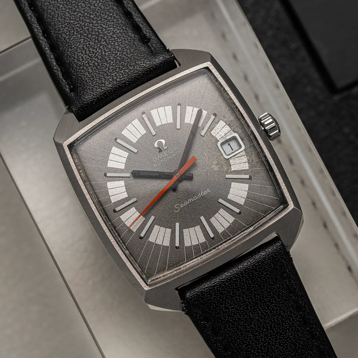 A vintage Omega Seamaster, Ref. 166.042, Cal. 565, Men’s Automaticwristwatch with a square stainless steel case, a gray dial featuring white hour markers and a red seconds hand, and a black leather strap, shown resting on a light gray textured surface.