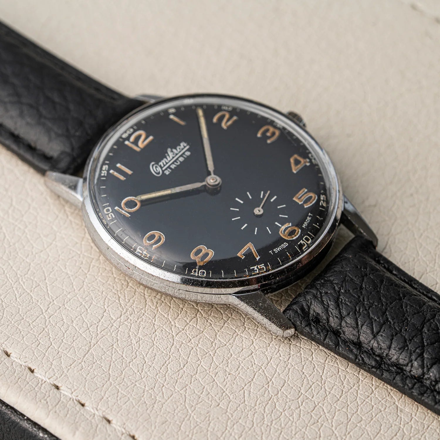 A vintage Omikron Ref. 3358, Cal. UT 6525 Swiss watch with a black dial, silver-toned case, and black leather strap, resting on a textured beige leather surface.