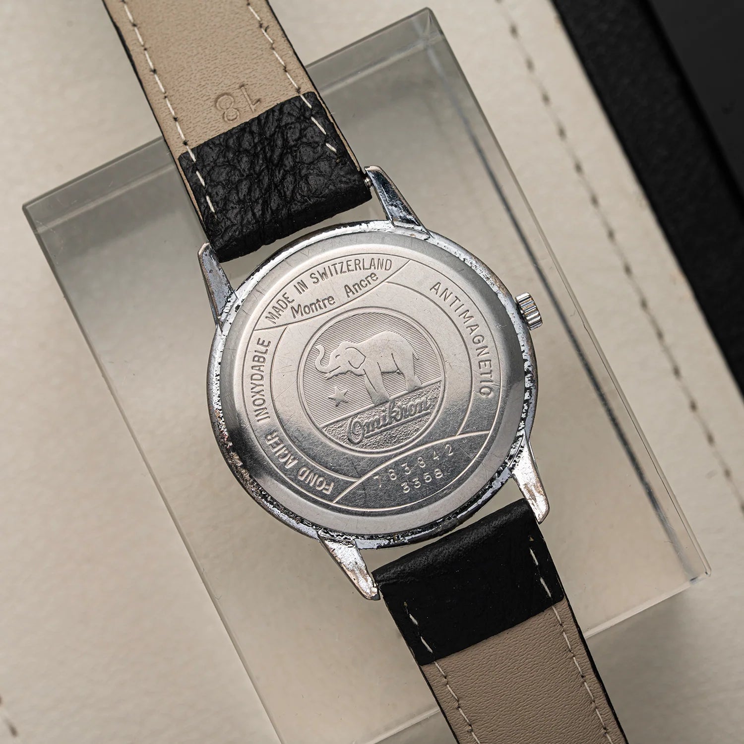 Close-up of the back of a vintage Omikron Ref. 3358, Cal. UT 6525 watch with a round silver-tone case, black and beige leather strap, and engraved details including the Cinnabar elephant logo and Montre-Aux-Chèvres text.