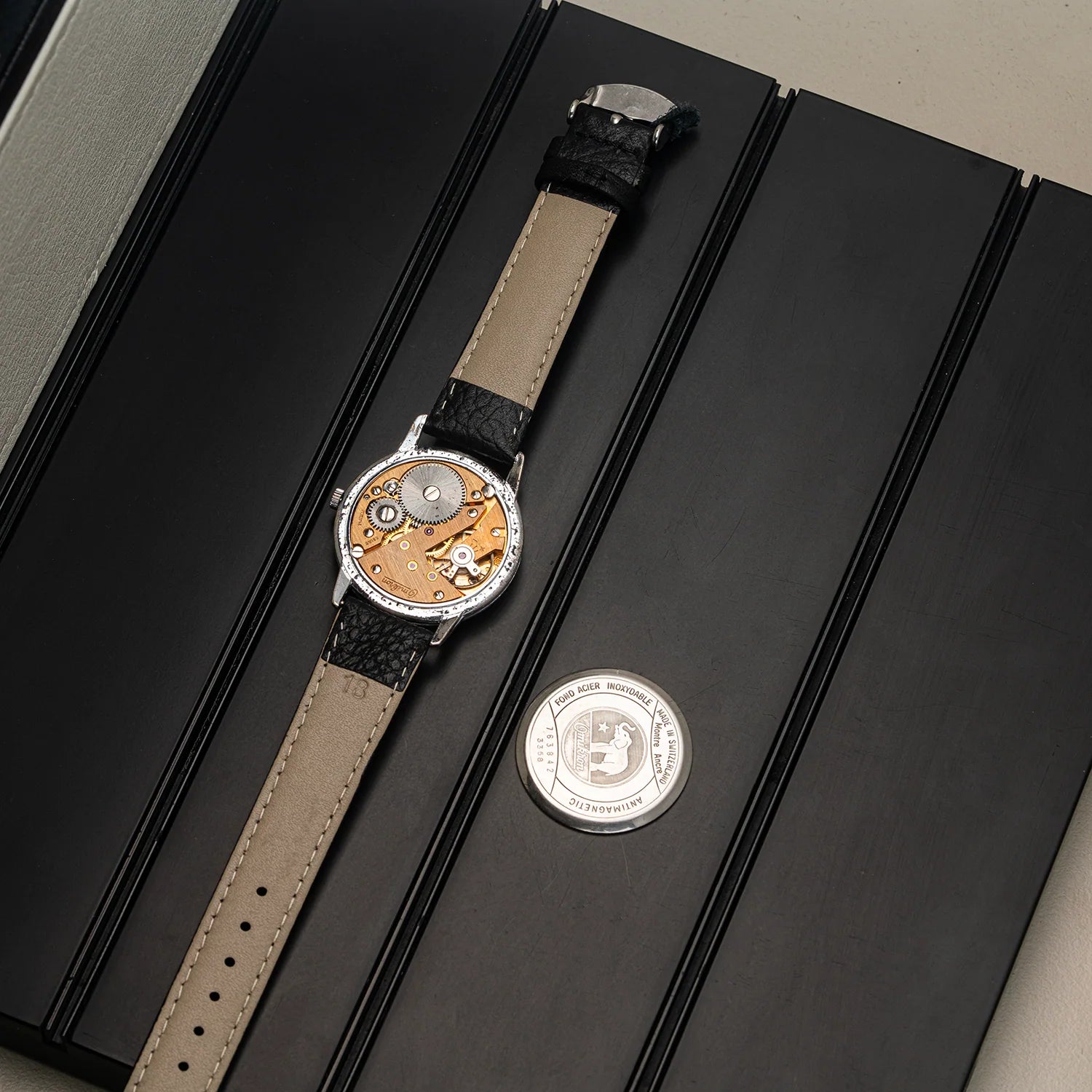 A luxury watch with a beige leather strap and a silver-toned case displaying a complex mechanical movement UT 6525, laid diagonally on a black presentation box next to a silver commemorative coin.