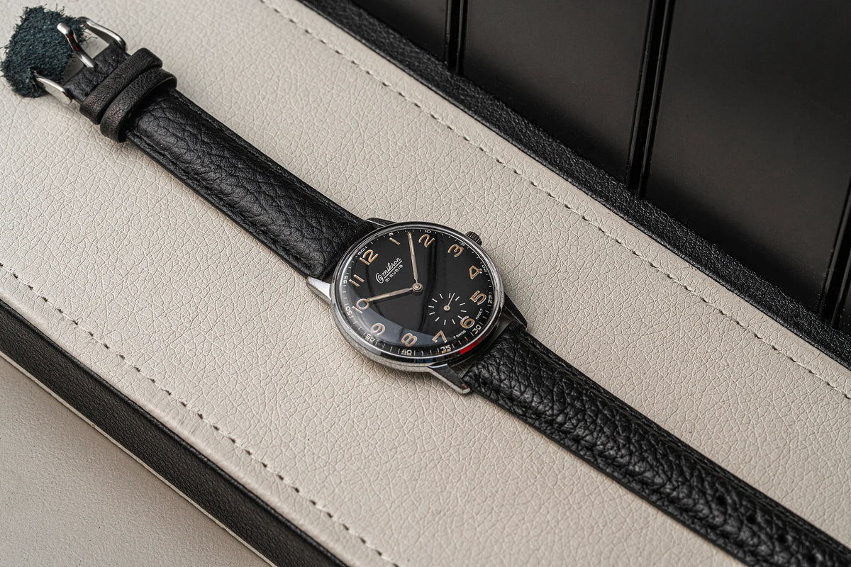 A wristwatch Omikron Ref. 3358, Cal. UT 6525 with a black textured leather strap and a black dial, resting diagonally on a cream-colored leather surface.