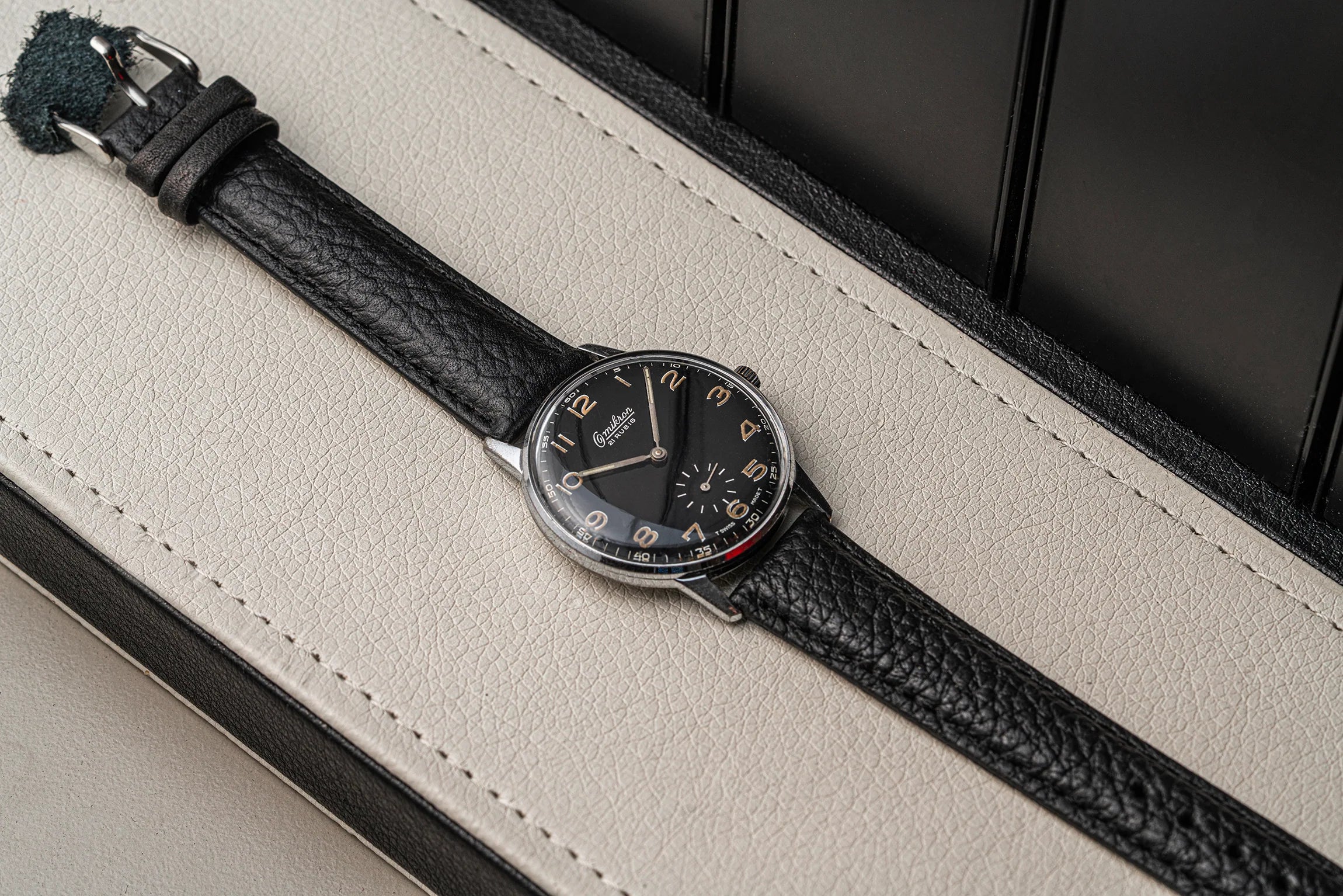 A wristwatch Omikron Ref. 3358, Cal. UT 6525 with a black textured leather strap and a black dial, resting diagonally on a cream-colored leather surface.