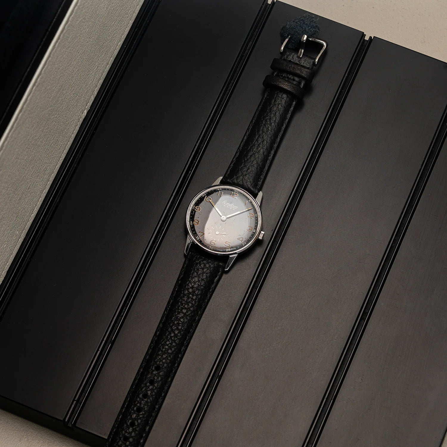 A wristwatch Omikron Ref. 3358, Cal. UT 6525 with a silver-toned case, white dial, and textured black leather strap, displayed diagonally on a dark gray presentation box.