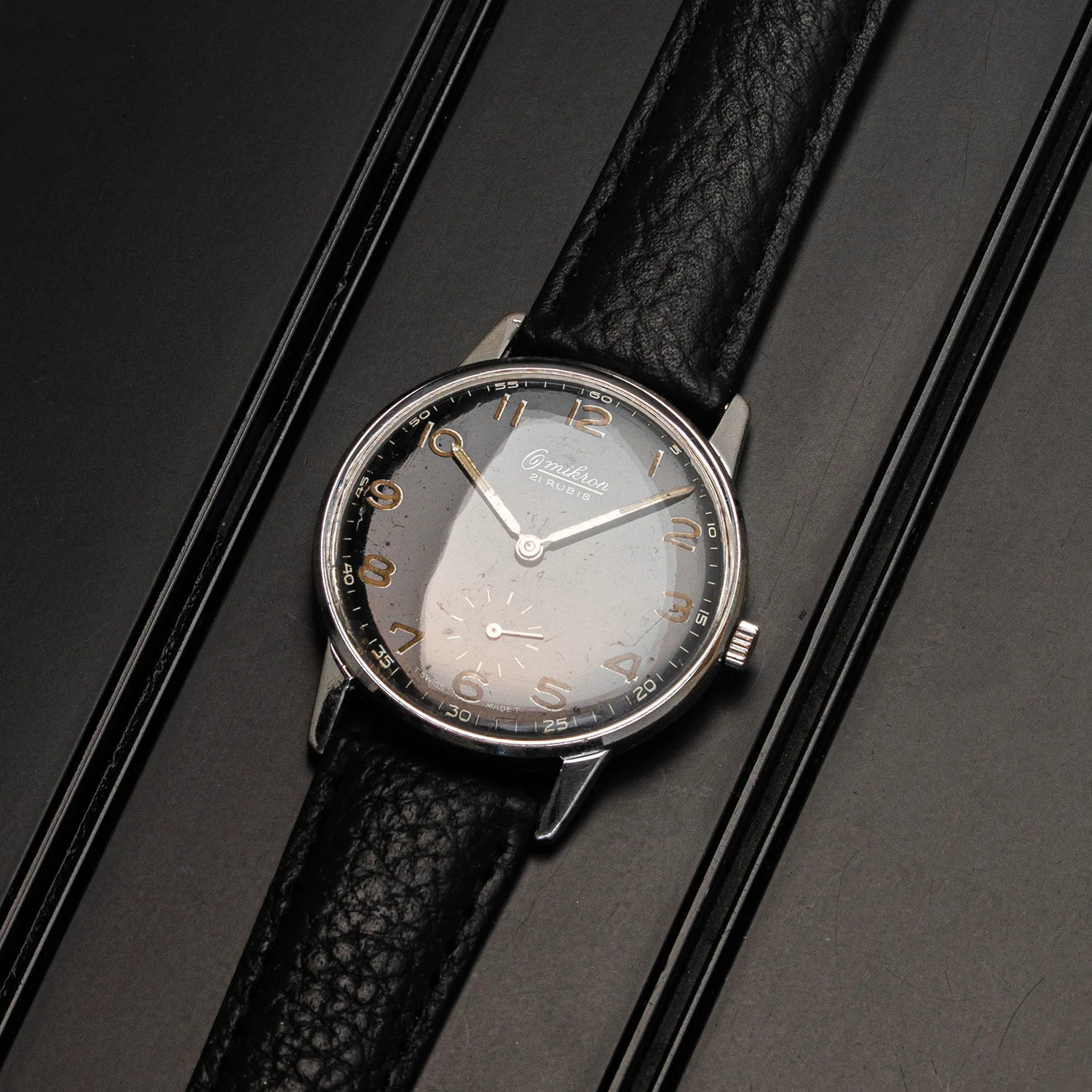 A vintage-style wristwatch Omikron Ref. 3358, Cal. UT 6525 with a silver-toned oval case, a light gray dial, and a black textured leather strap, photographed on a dark gray background with diagonal black bars.