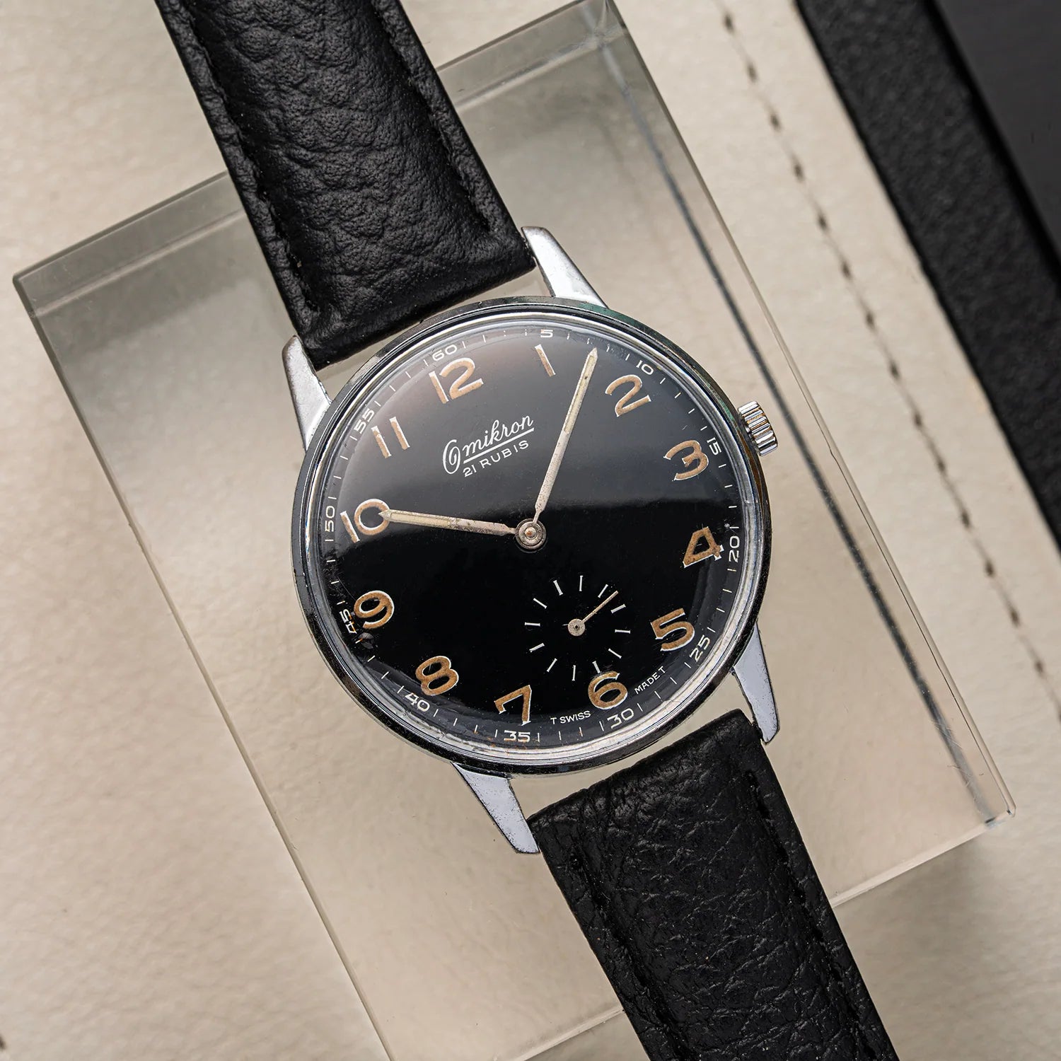 A vintage-style wristwatch Omikron Ref. 3358, Cal. UT 6525 with a black dial, silver-toned case, and black leather strap, displayed diagonally on a clear acrylic block over a beige textured surface.