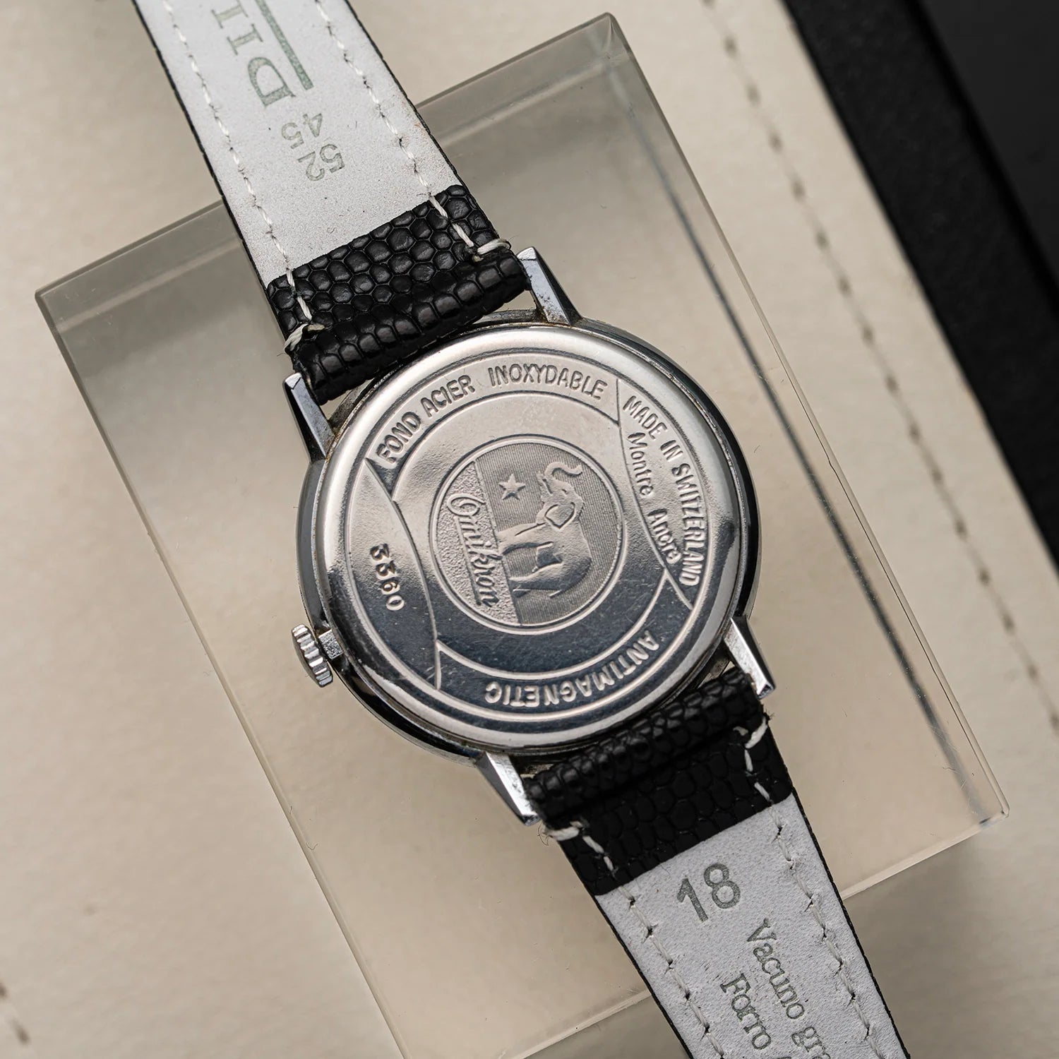 A close-up of a vintage wristwatch Omikron, Ref. 3360, Cal. AS 1130 shown from the back, featuring a round silver-tone case with engraved text and emblem, and a black-and-white patterned strap, resting on a clear display box.