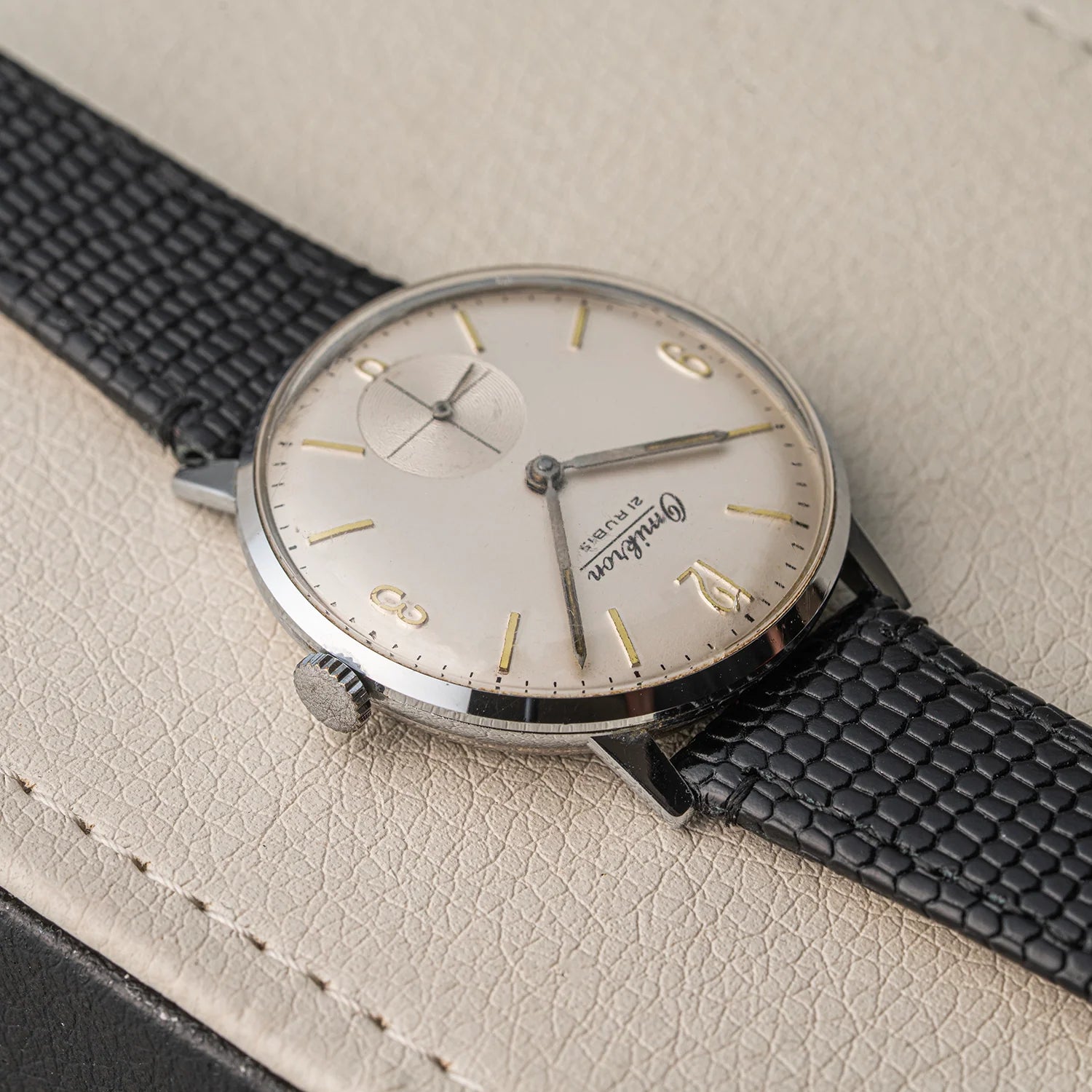 A vintage-style wristwatch Omikron, Ref. 3360, Cal. AS 1130 with a silver-toned oval case, cream dial, gold-tone hour markers and hands, and a black textured leather strap, photographed on a light beige leather surface.