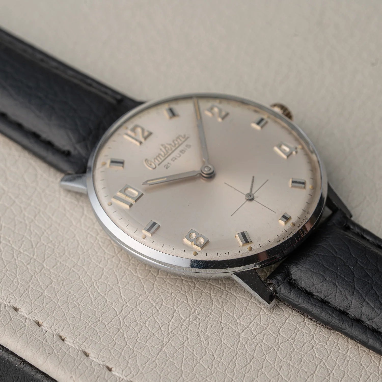 A vintage Omikron AS 1130 watch with a silver-toned round case, cream-colored dial, and black leather strap, resting on a textured light beige leather surface.