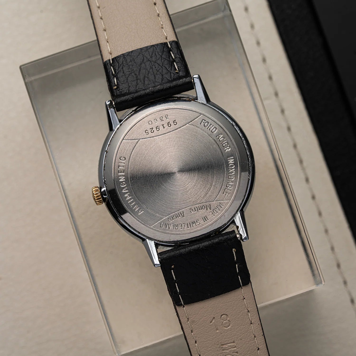 Close-up of the back of a wristwatch Omikron AS 1130 with a brushed silver-tone case and a black-and-cream leather strap, resting on a clear display box.