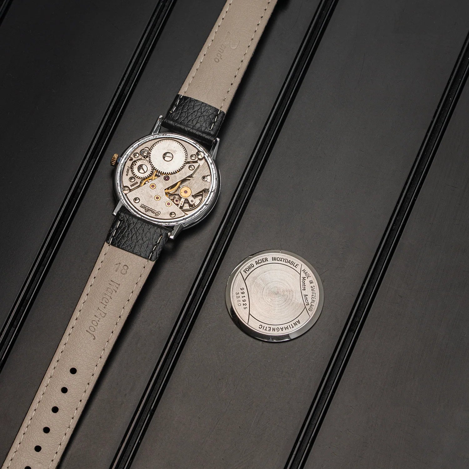 A vintage watch Omikron AS 1130 showin the internal mechanical movement, paired with a light beige leather strap, displayed diagonally on a dark textured background next to its circular metal case lid.