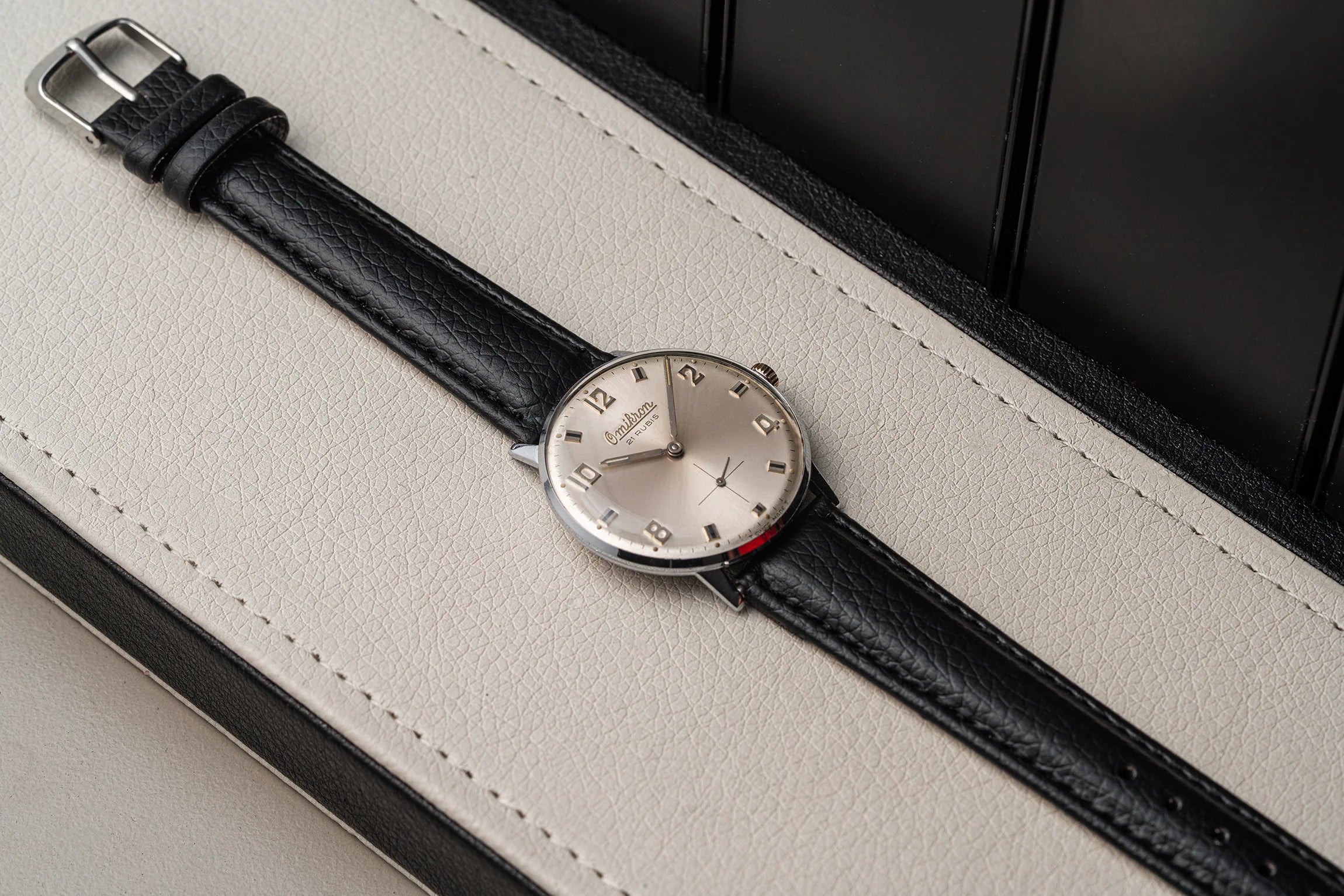 A vintage-style wristwatch Omikron AS 1130 with a black leather strap and a round silver-toned dial, resting diagonally on a light beige textured leather surface with a dark background.