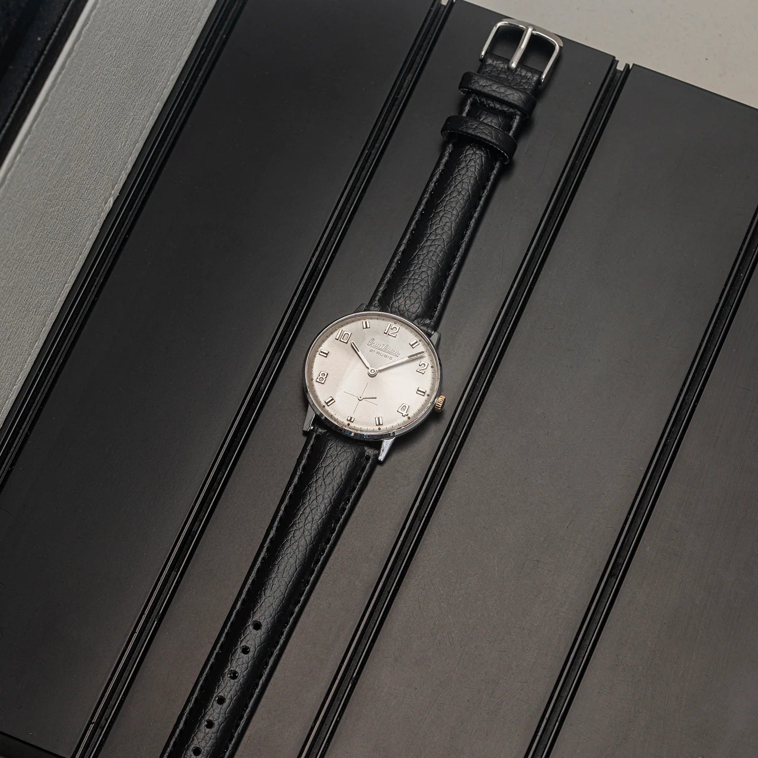A vintage-style wristwatch Omikron AS 1130 with a black textured leather strap and a silver-toned round dial, displayed diagonally on a dark gray textured surface.