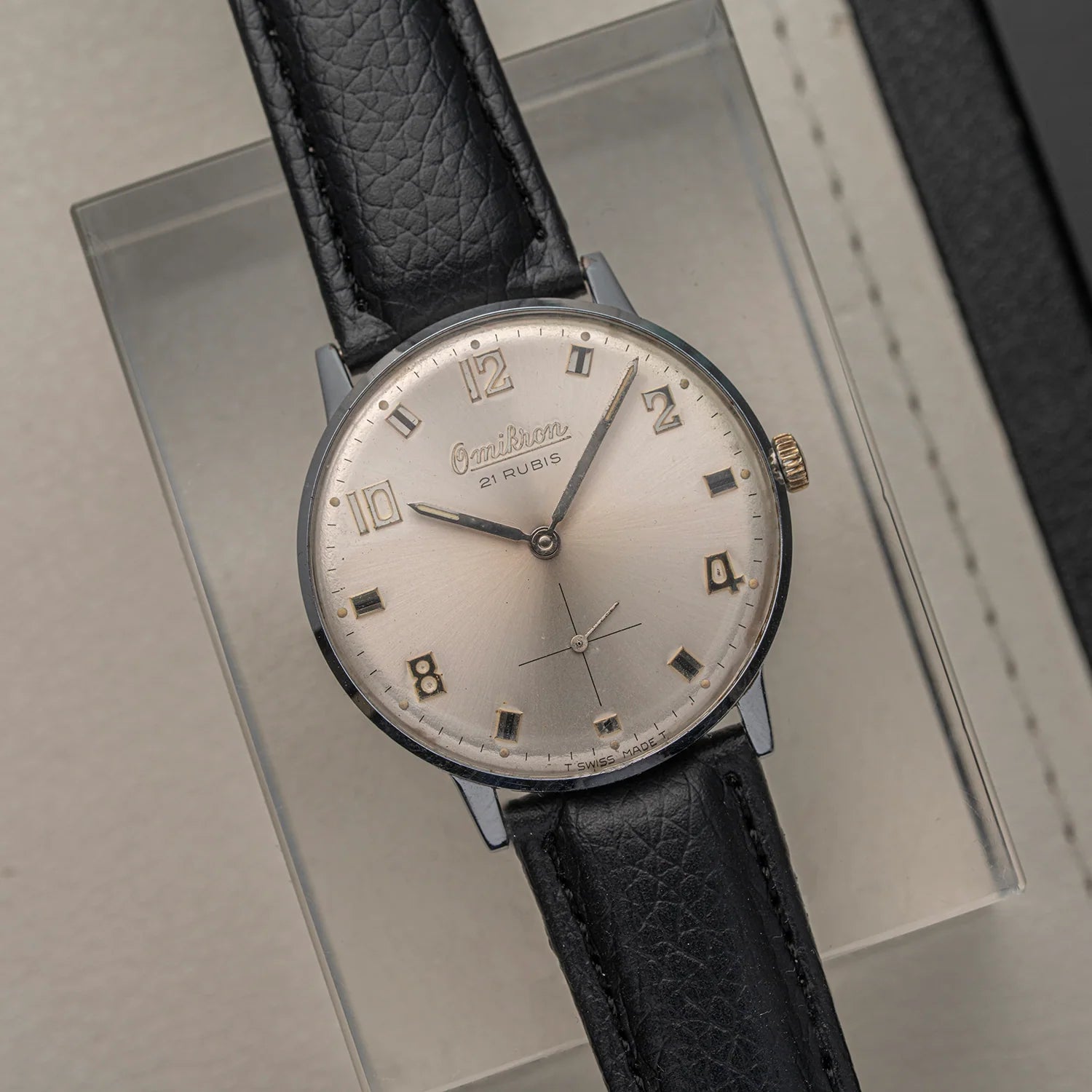 A vintage-style wristwatch Omikron AS 1130 with a round silver-toned case, a pale champagne dial, and a black textured leather strap, displayed diagonally on a clear acrylic stand.