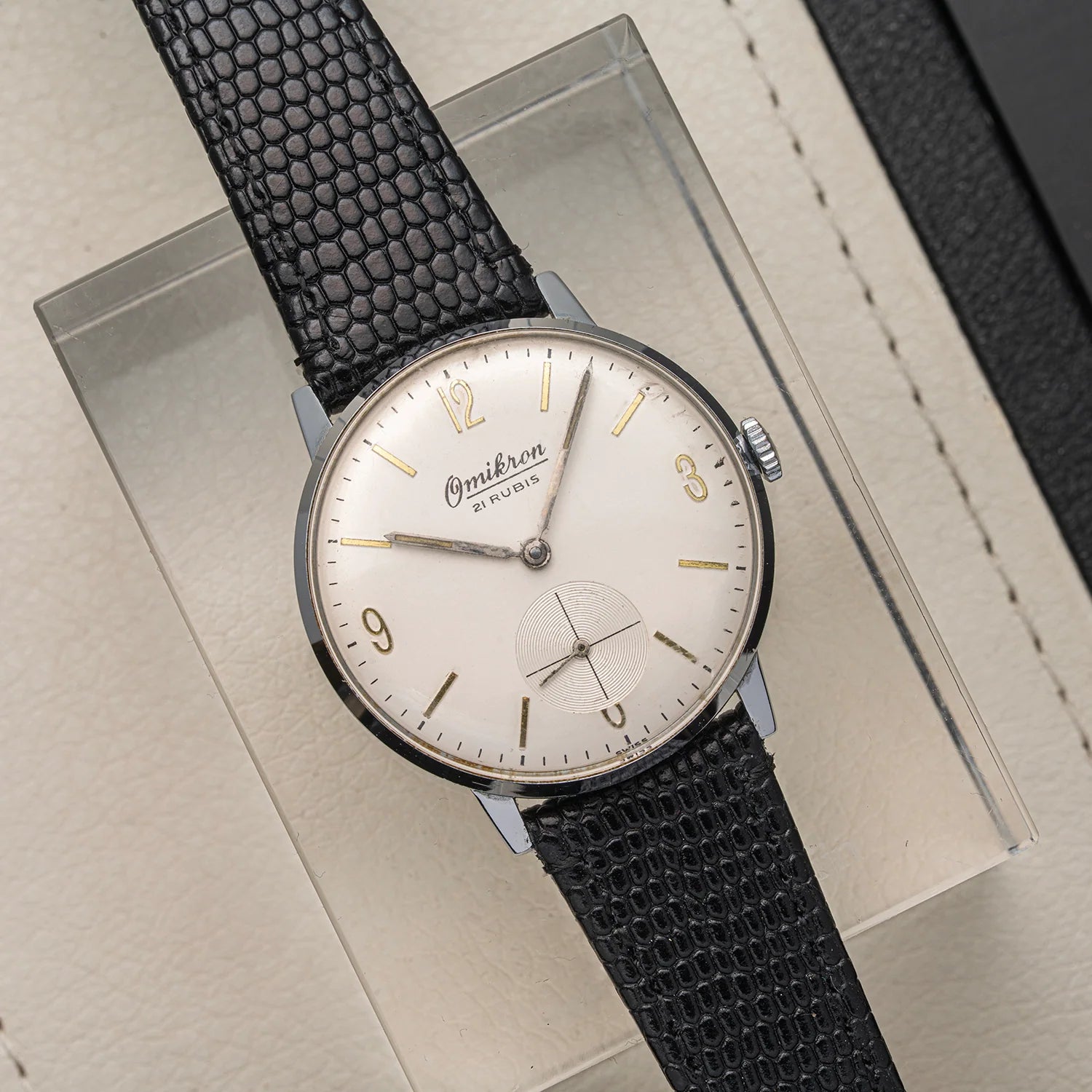 An Omikron, Ref. 3360, Cal. AS 1130 wristwatch with a silver-toned oval case, cream dial, and textured black strap, displayed on a clear acrylic stand over a beige surface.