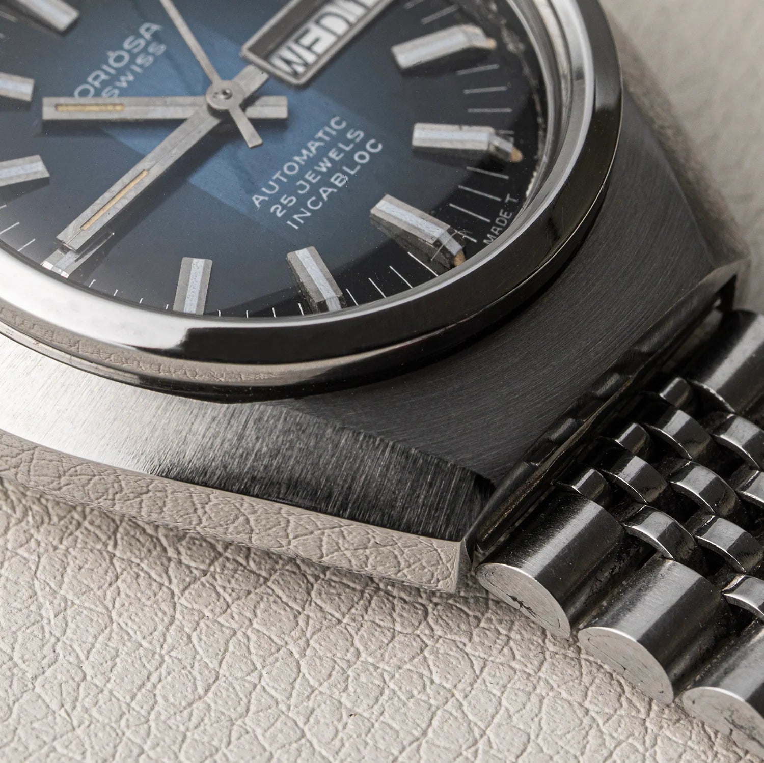 Close-up of an Oriosa, ETA 2789 Swiss Automatic 25 Jewels Incabloc watch with a dark blue dial, brushed stainless steel case, and matching metal bracelet resting on a textured light beige surface.