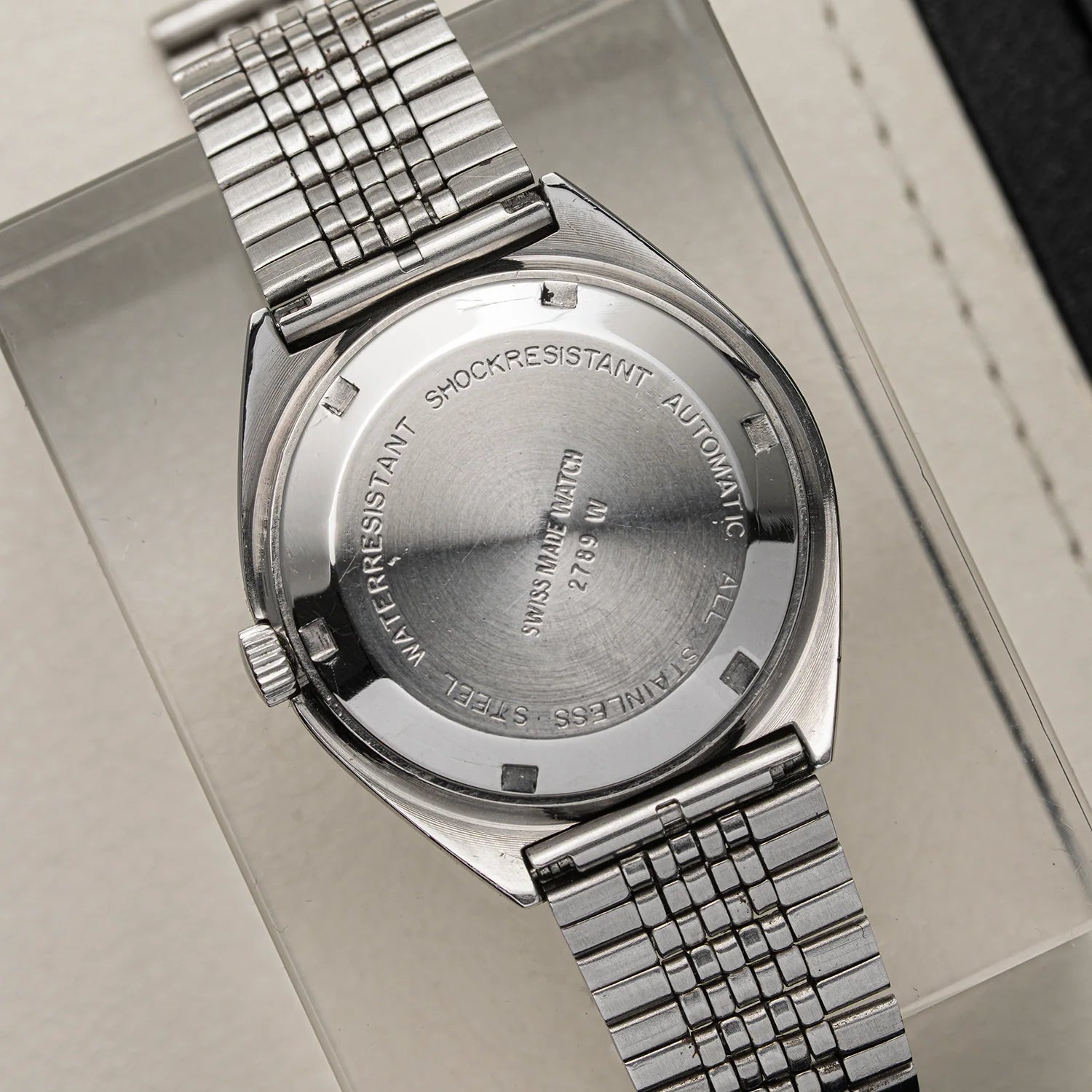 Close-up of the back of a stainless steel wristwatch Oriosa, ETA 2789 with a metal link bracelet, resting on a light beige surface. The case is engraved with text including 'SWISS MADE 31L STEEL' and 'SHOCKRESISTANT AUTOMATIC'.