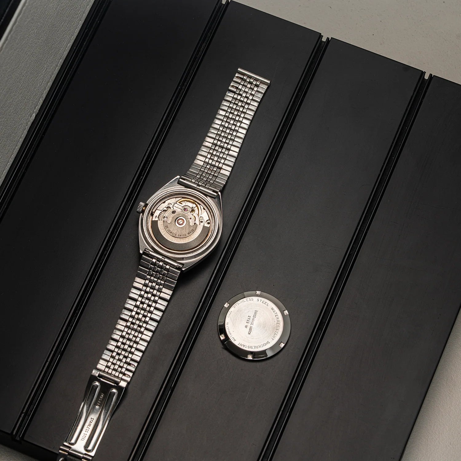 A silver stainless steel automatic watch  Oriosa, ETA 2789with a metal link bracelet, shown from the back with the case open, resting on a black presentation box next to a small round metal component.