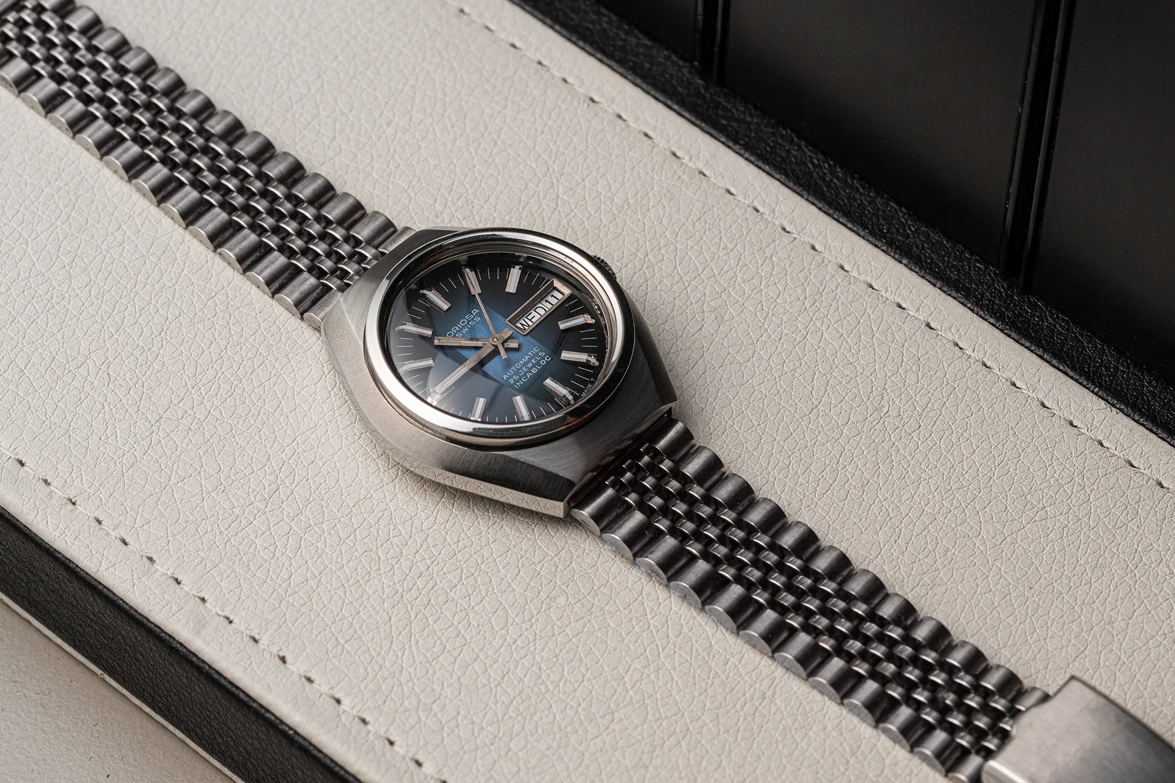 A close-up of a wristwatch Oriosa, ETA 2789 with a dark blue dial and a silver-toned metal link bracelet, resting on a light textured leather surface.