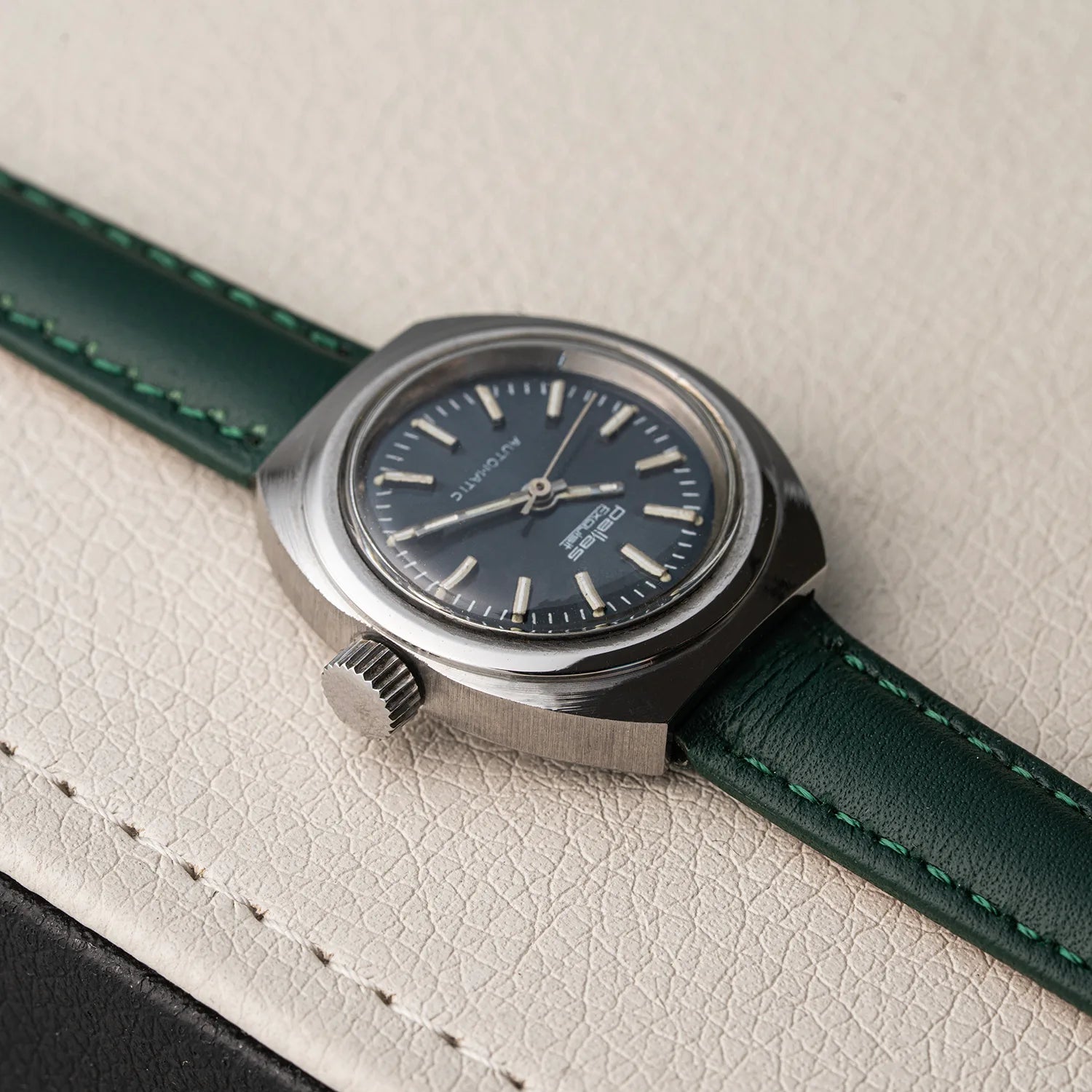 A vintage-style wristwatch Pallas Exquisit ETA 2670 with a dark blue dial, silver-toned metal case, and a dark green leather strap, resting on a textured off-white leather surface.