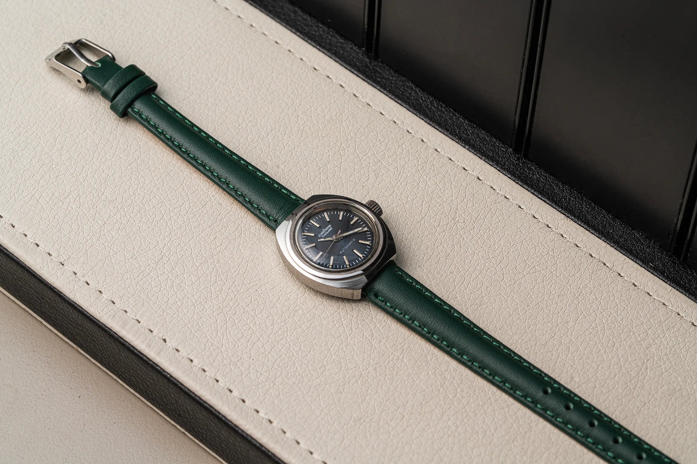 A wristwatch Pallas Exquisit ETA 2670 with a dark green leather strap and a silver-toned round case, resting diagonally on a cream-colored textured leather surface.