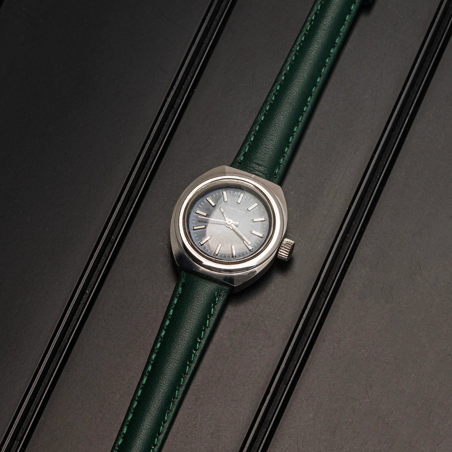 A vintage-style wristwatch Pallas Exquisit ETA 2670 with a round silver-tone case, a light gray dial, and a dark green leather strap with matching green stitching, displayed diagonally on a dark textured background.