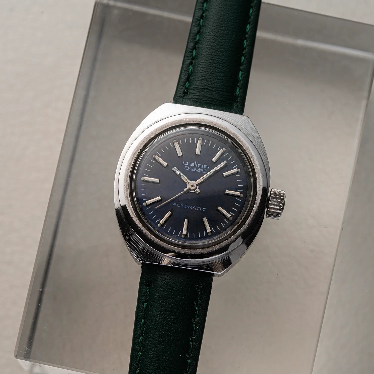 A vintage-style wristwatch Pallas Exquisit ETA 2670 with a silver-toned round case, dark blue dial, and dark green leather strap, displayed on a clear acrylic stand against a light gray background.