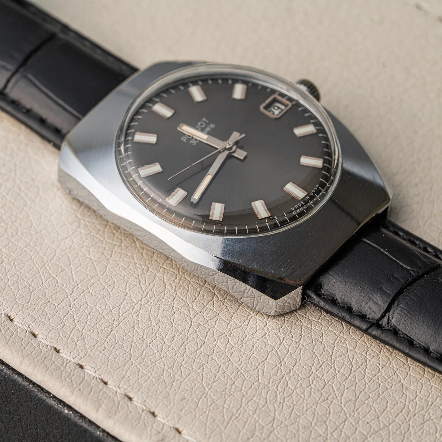 A vintage-style wristwatch Poljot 30 Jewels with a dark gray dial, silver-toned case, and black leather strap, resting on a textured beige leather surface.