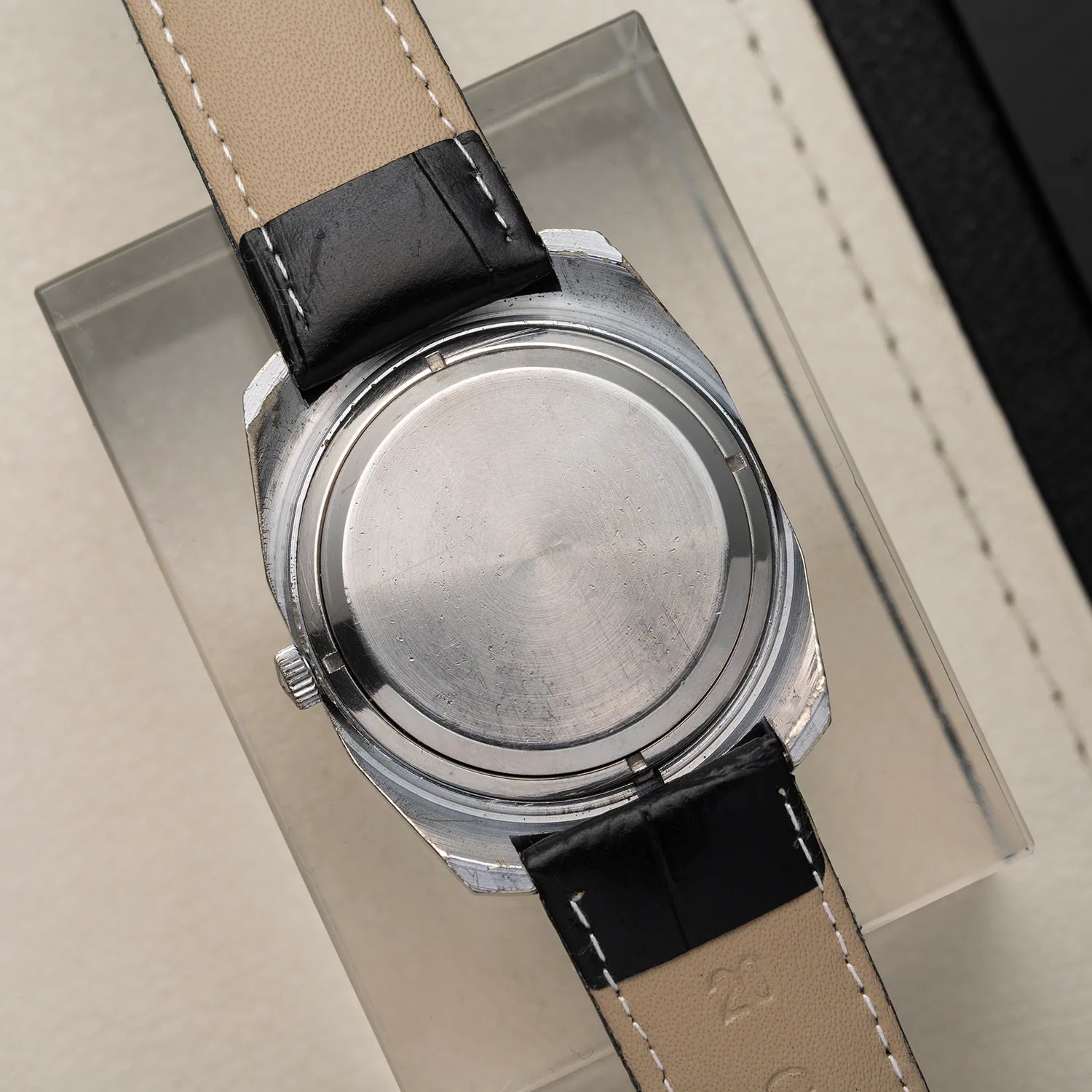 Close-up of the back of a vintage wristwatch Poljot 30 Jewels with a brushed silver metal case and a black-and-cream leather strap, resting on a clear display block.