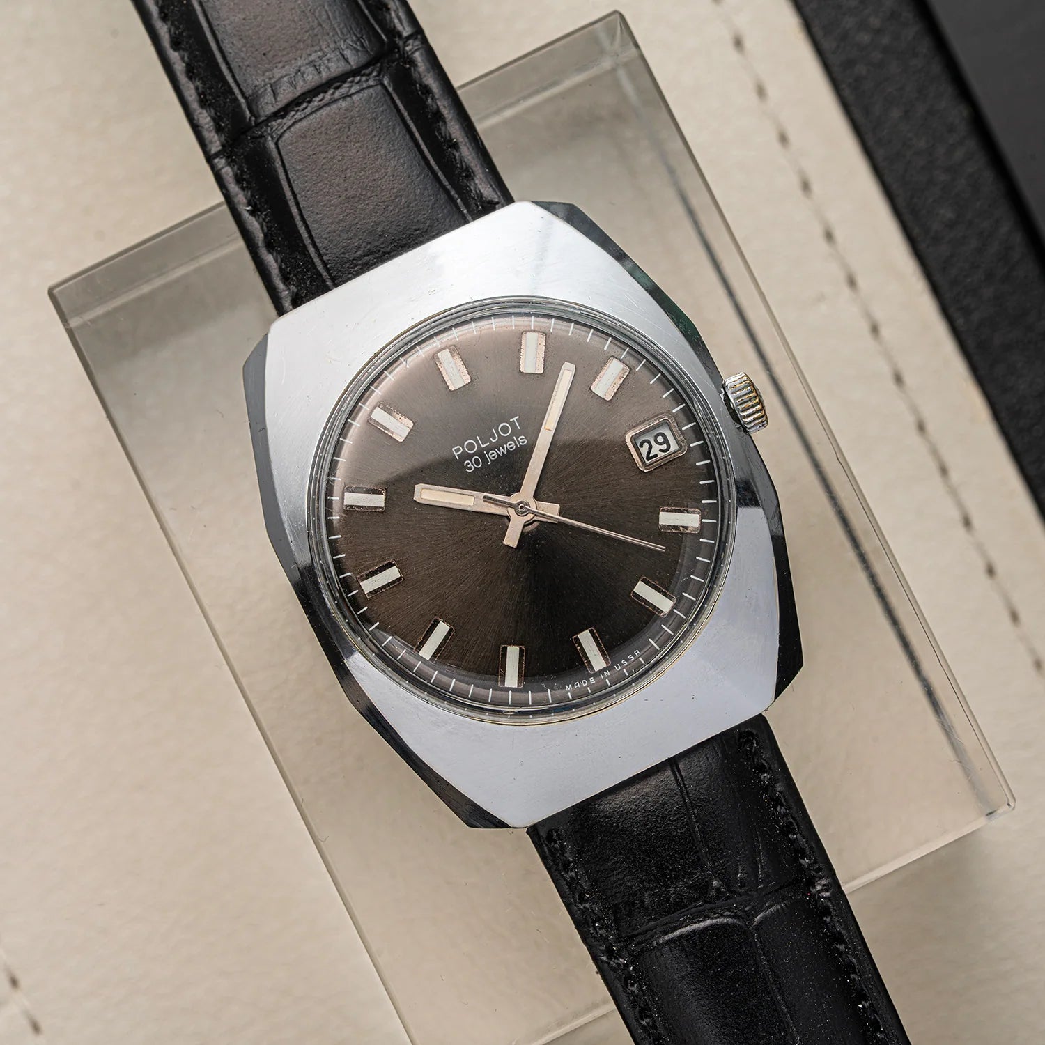 A vintage-style wristwatch Poljot 30 Jewels with a silver-toned metal case, dark brown dial, and black leather strap, displayed on a clear acrylic stand against a beige textured background.