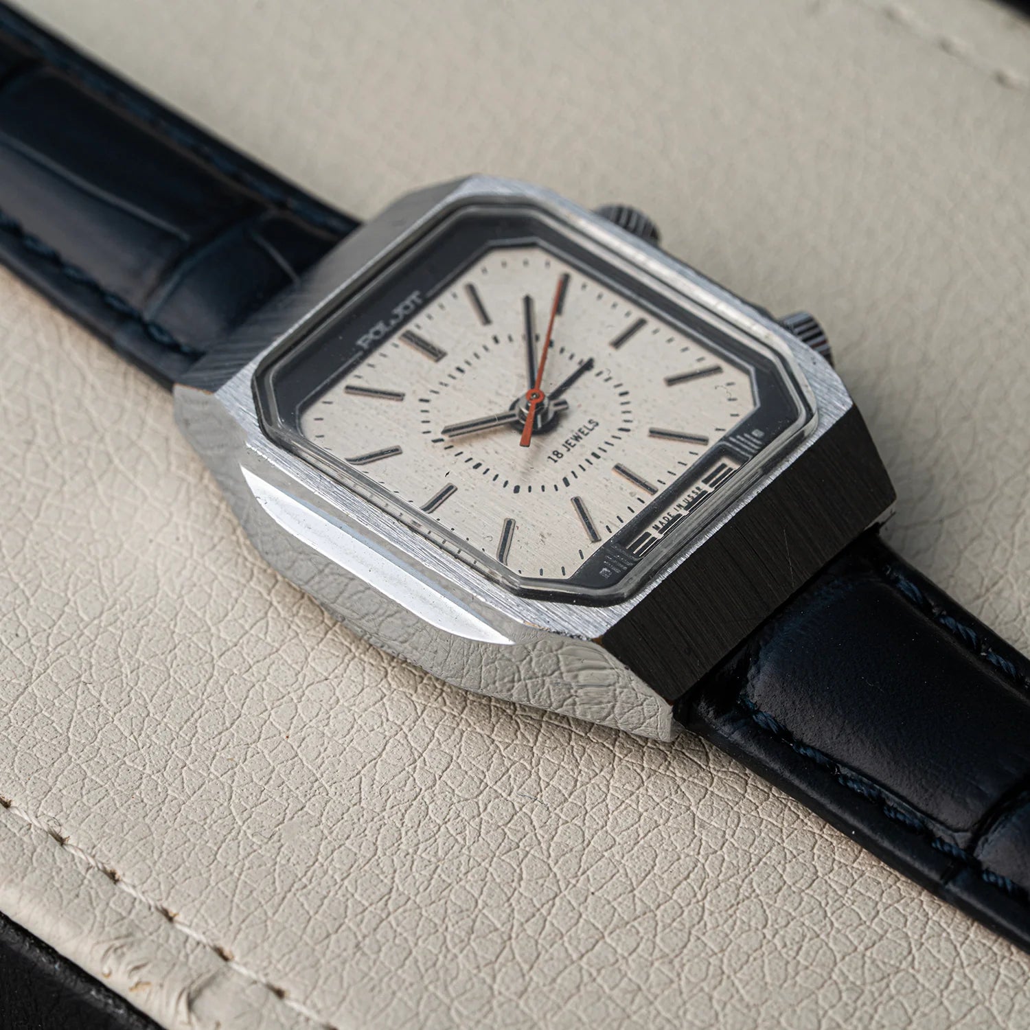 A close-up of a wristwatch Poljot Alarm, Cal. 2612.1 with a brushed silver-tone case, white dial, and dark navy leather strap, resting on a textured beige surface.