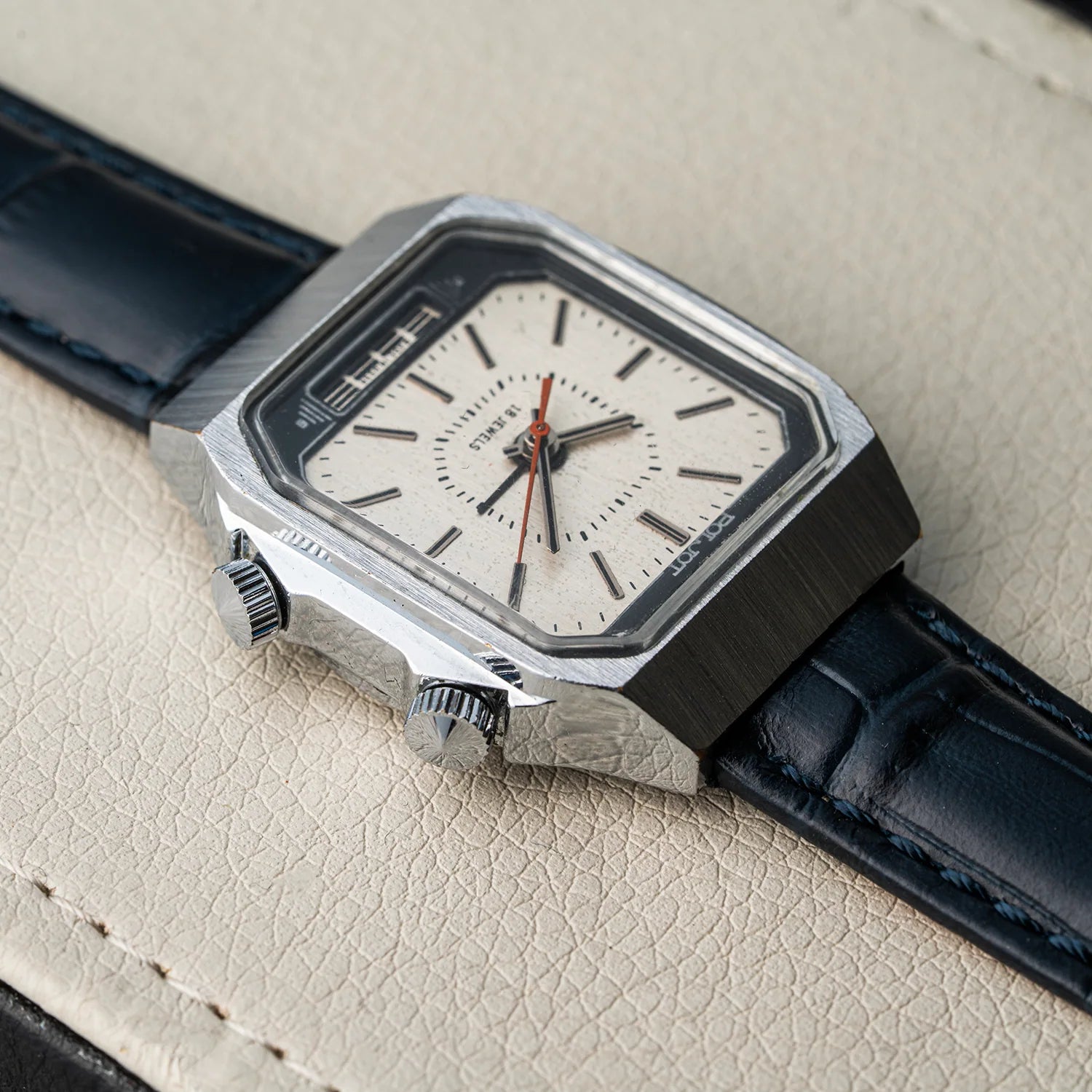 A close-up of a wristwatch Poljot Alarm, Cal. 2612.1 with a silver-toned metal case, a white dial, and a dark blue leather strap, resting on a textured beige surface.