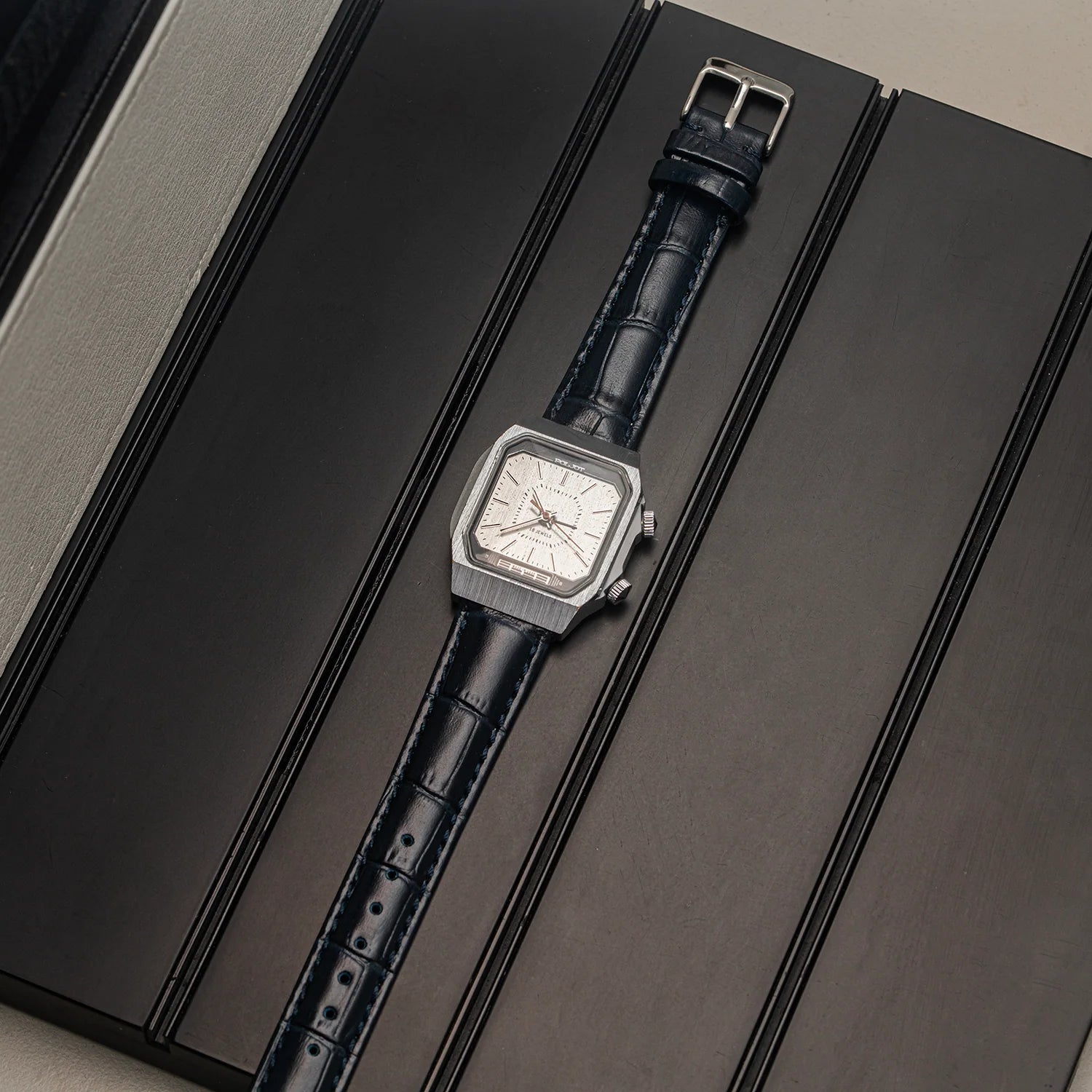 A rectangular silver-tone wristwatch Poljot Alarm, Cal. 2612.1 with a white dial and a dark blue leather strap, displayed diagonally on a dark textured presentation box.