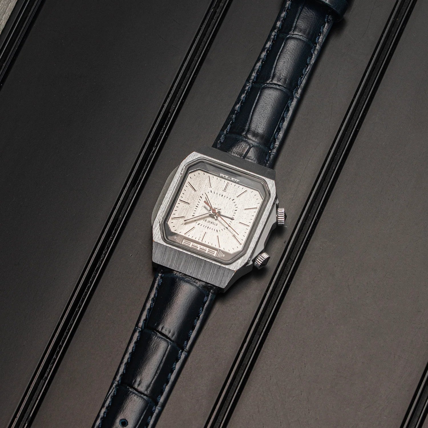 A vintage-style wristwatch Poljot Alarm, Cal. 2612.1 with a brushed silver-tone octagonal case, white dial, and dark blue leather strap, photographed diagonally on a dark textured background.