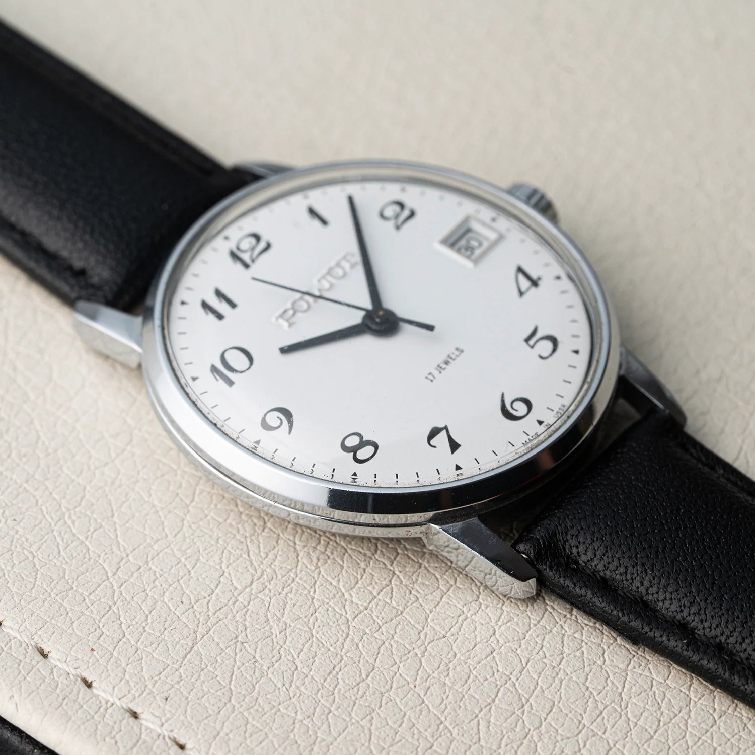 A close-up of a vintage-style wristwatch Poljot 2614.2H with a white dial, black Arabic numerals, black hands, and a black leather strap, resting on a textured beige surface.