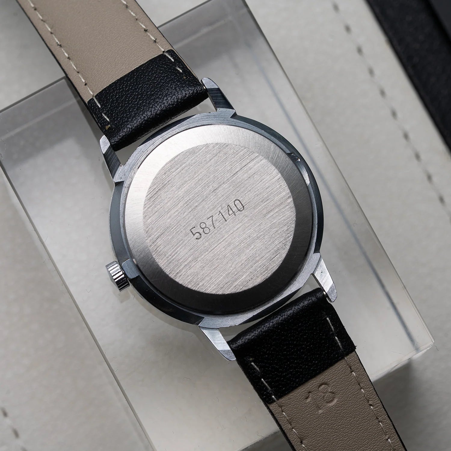 Close-up of the back of a wristwatch Poljot 2614.2H with a brushed silver-tone case and a two-tone strap in beige and black leather, resting on a light gray box.