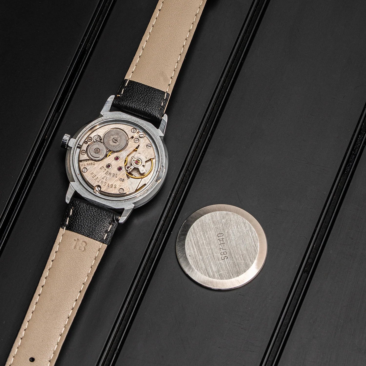 A close-up of a vintage wristwatch Poljot 2614.2H with a silver-toned case, beige leather strap, and visible mechanical movement, placed on a black textured surface next to a round brushed metal tag.