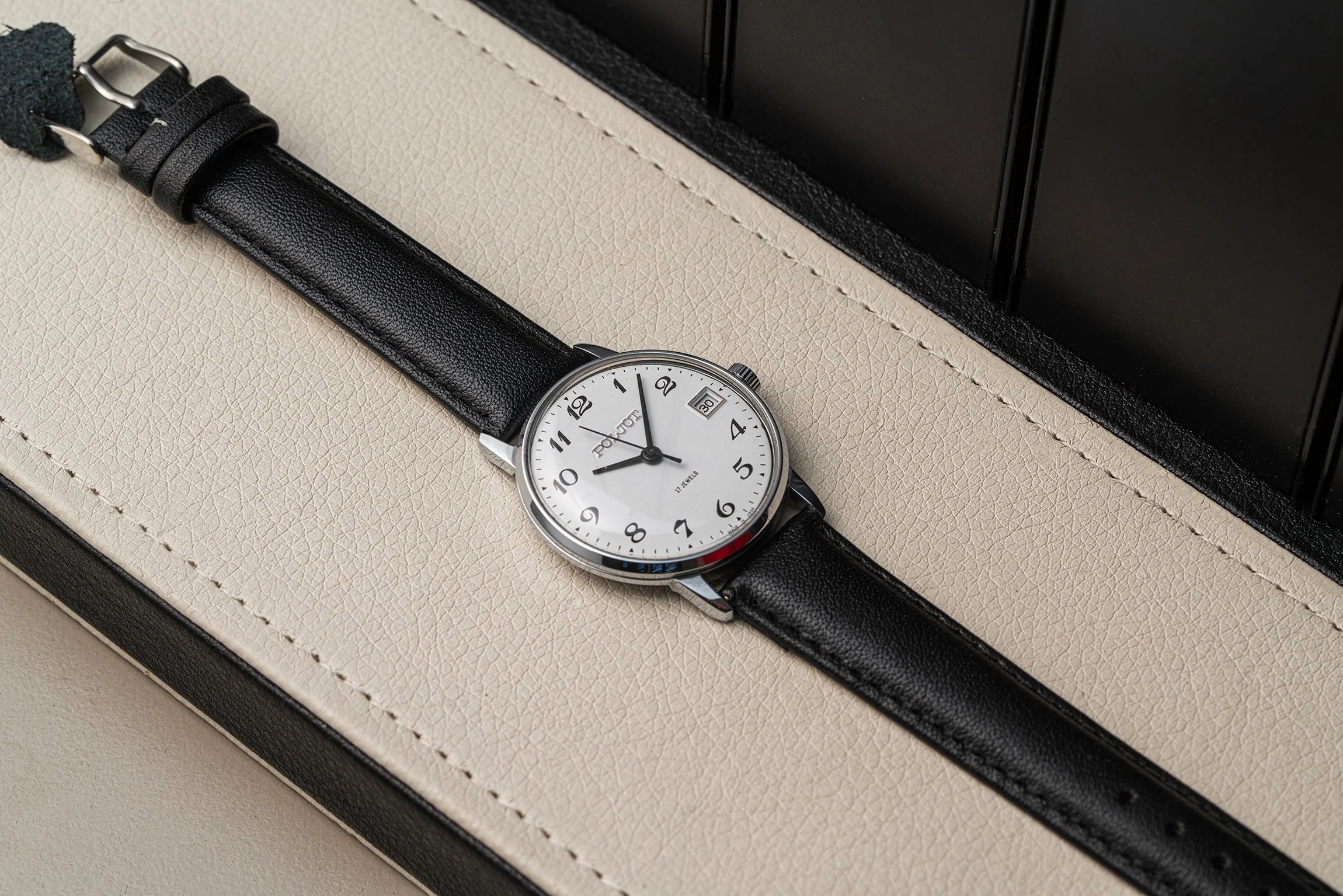 A wristwatch Poljot 2614.2H with a black leather strap and a silver-toned case, featuring a white dial with black Arabic numerals and a date window, resting on a cream-colored textured surface.