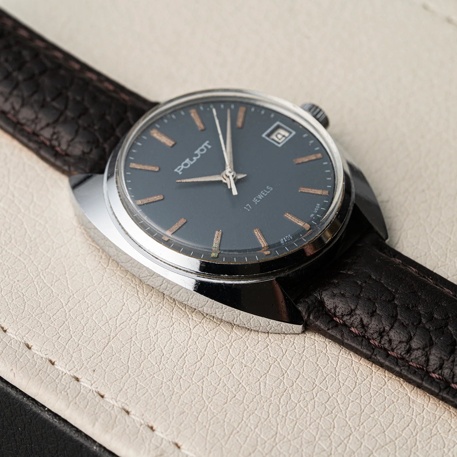 A close-up of a Poljot, Cal. 2614.2H watch with a dark gray dial, silver-tone case, and a textured dark brown leather strap, resting on a cream-colored textured surface.
