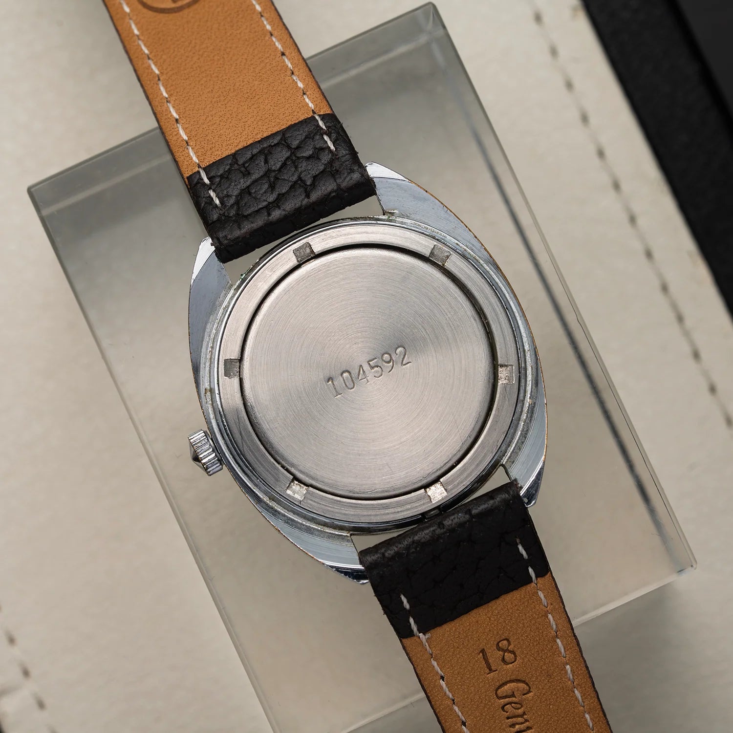 Close-up of the back of a wristwatch Poljot, Cal. 2614.2H with a round silver-tone case and a tan leather strap with black textured sections, resting on a clear display block.