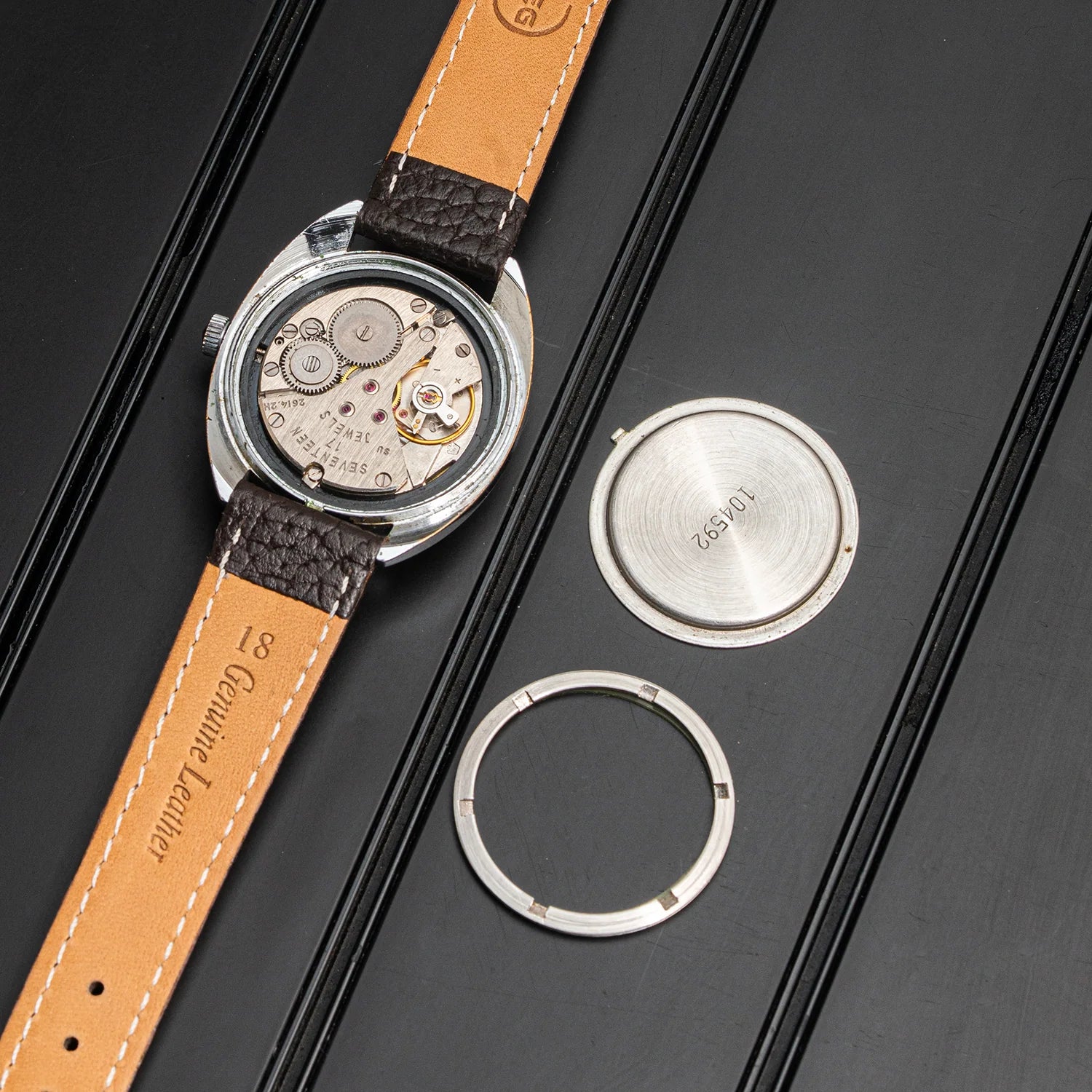 A vintage mechanical watch Poljot, Cal. 2614.2H with a tan leather strap and silver-toned case, shown alongside its separate silver back cover and ring on a black textured background. The strap is embossed with 'Genuine Leather' and 'W.