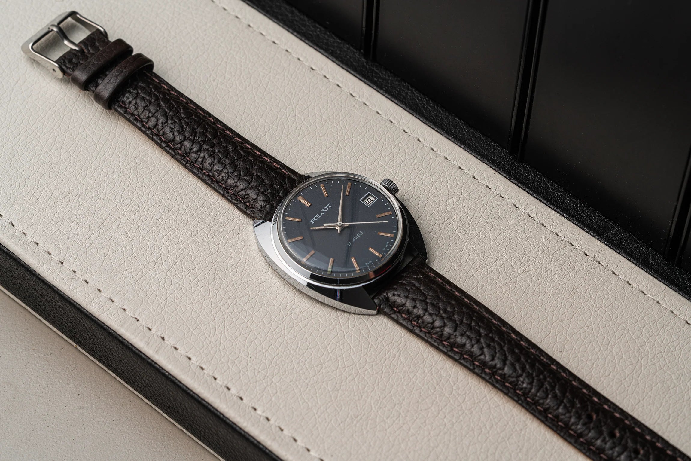 A close-up of a wristwatch Poljot, Cal. 2614.2H with a dark textured leather strap and a dark dial, resting diagonally on a cream-colored leather surface with a black background.