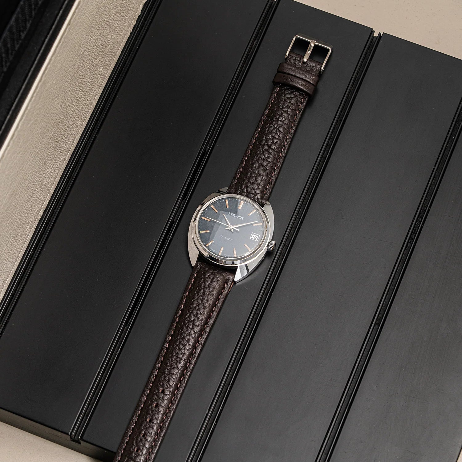 A wristwatch Poljot, Cal. 2614.2H with a dark brown textured leather strap and a silver-toned round case, shown resting diagonally on a black presentation box.