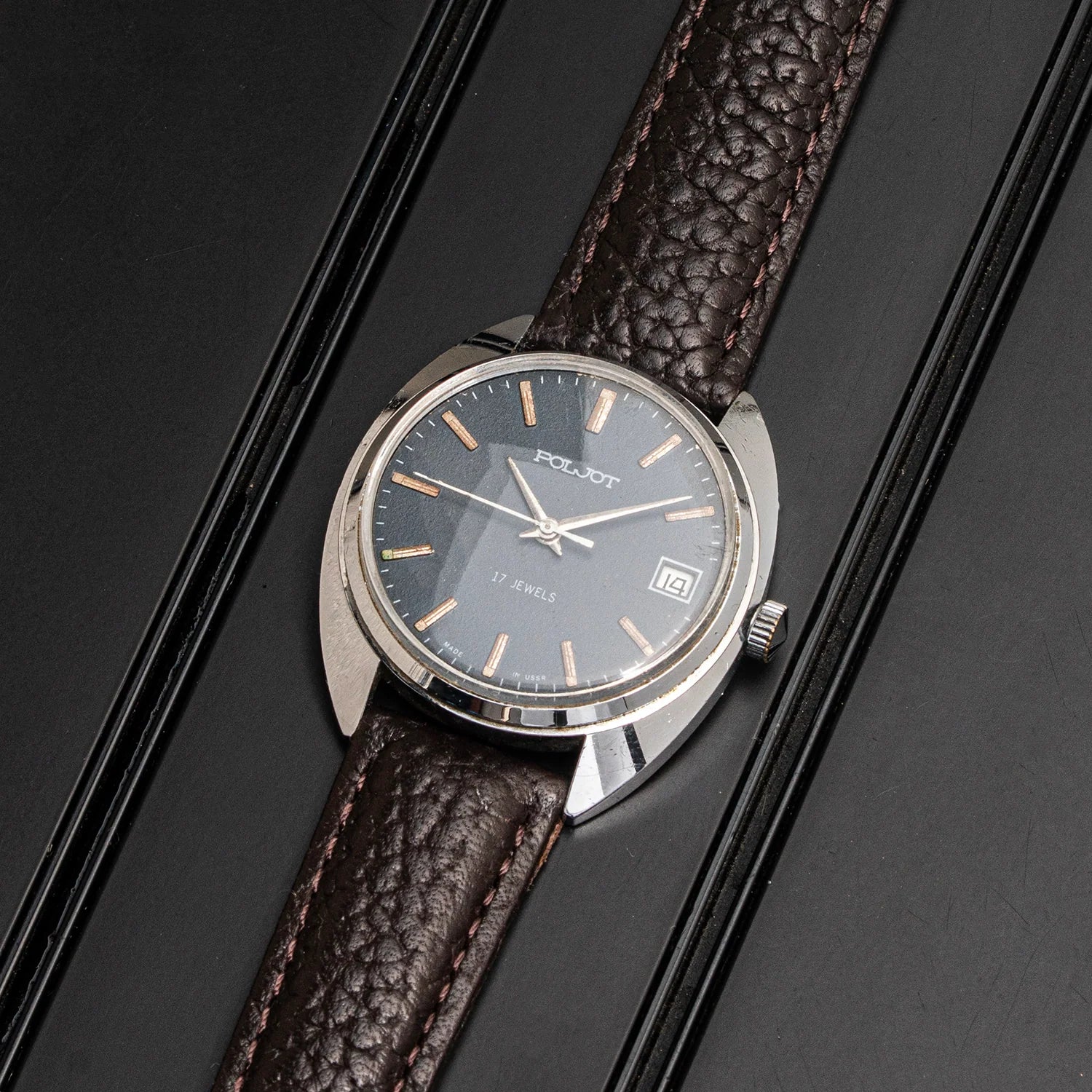 A vintage-style wristwatch Poljot, Cal. 2614.2H with a silver-toned round case, dark gray dial, and brown textured leather strap, photographed diagonally on a black background.