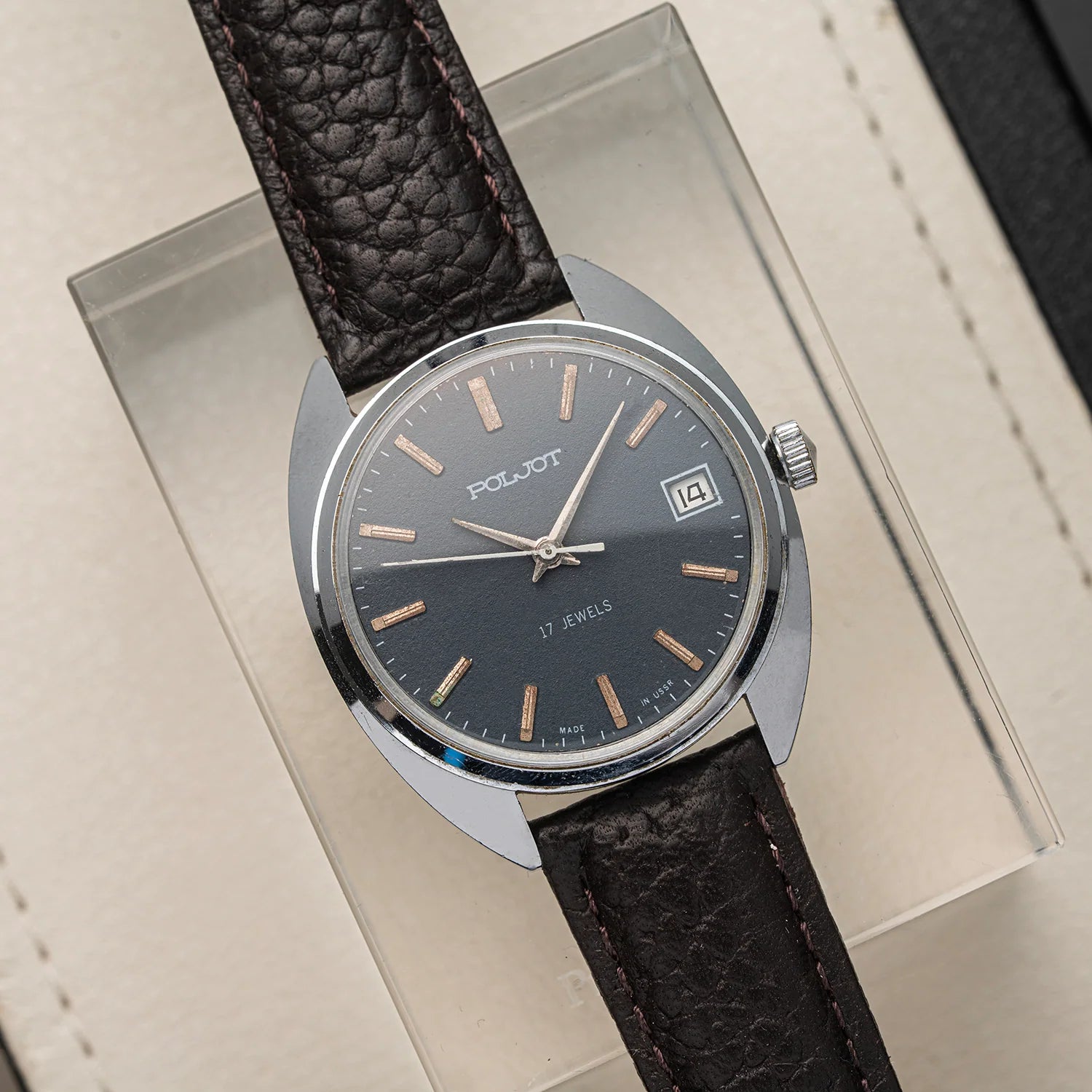 A vintage Poljot, Cal. 2614.2H wristwatch with a dark gray dial, silver-tone case, and textured dark brown leather strap, resting on a clear acrylic block over a beige surface.
