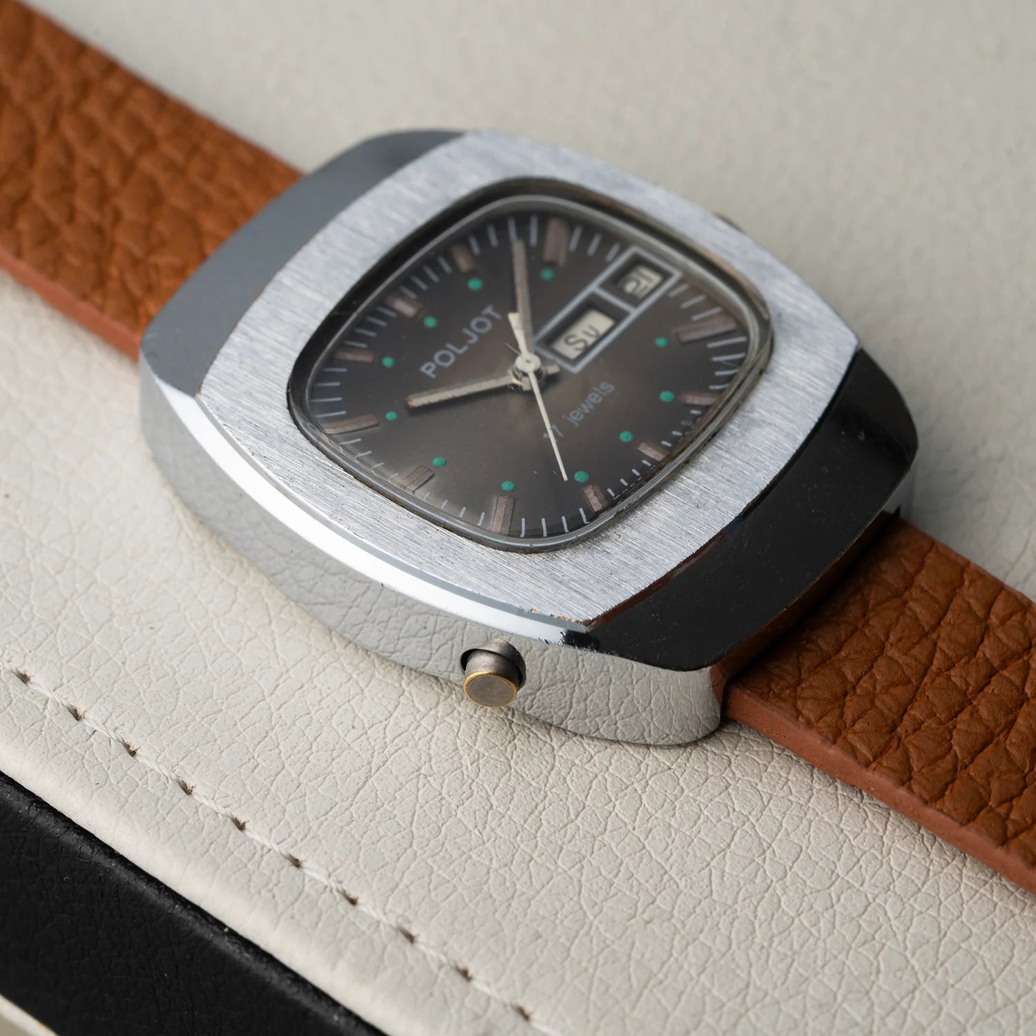 A vintage Poljot wristwatch with a silver-toned metal case, dark dial, and brown textured leather strap, resting on a light beige textured surface.