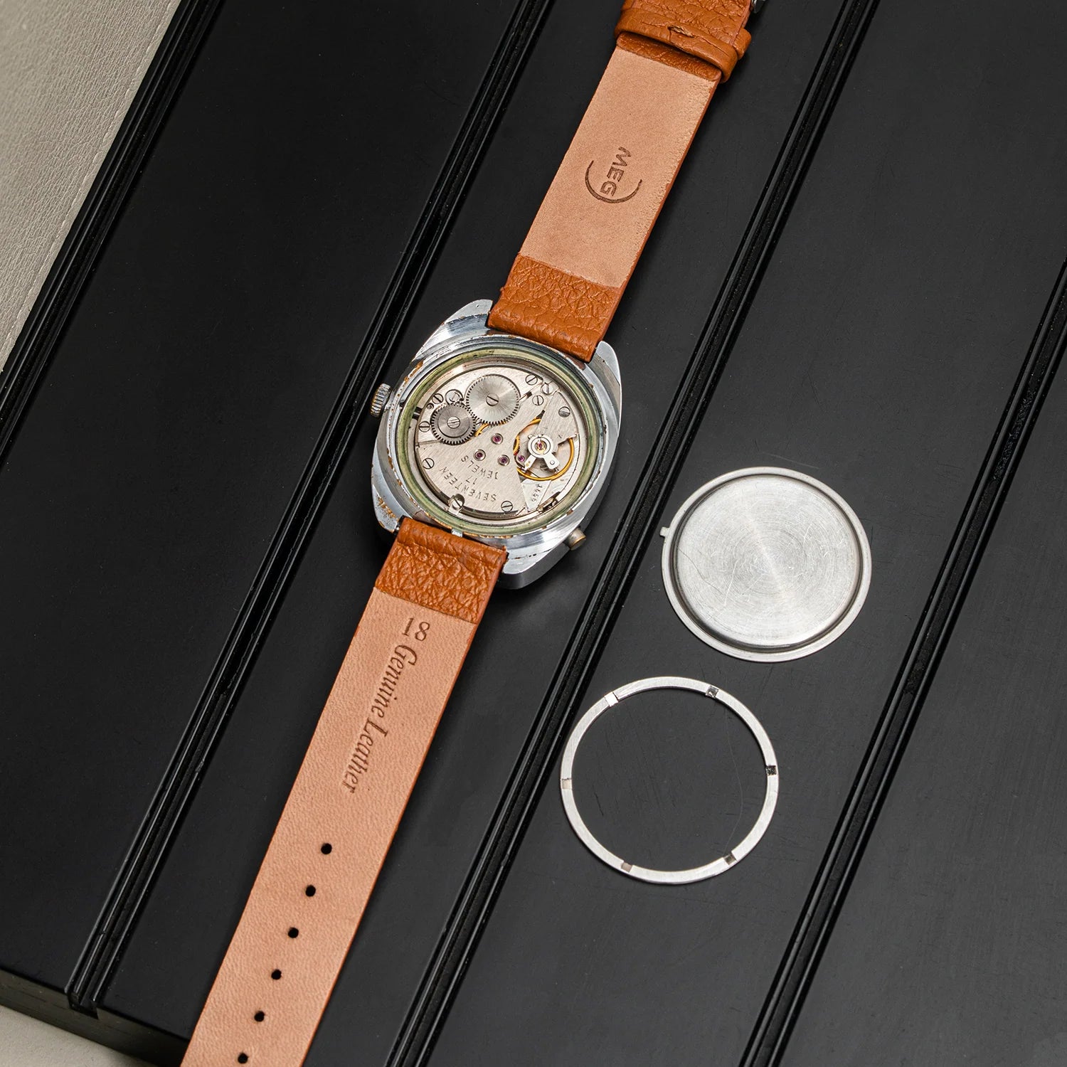 A vintage-style wristwatch with a tan leather strap and exposed mechanical movement, laid diagonally on a black presentation box alongside two silver watch parts.